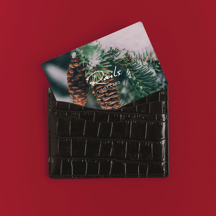 Rails gift card with a snowy pine cone and evergreen branch design, tucked into a dark brown croc-embossed leather cardholder on a red background.