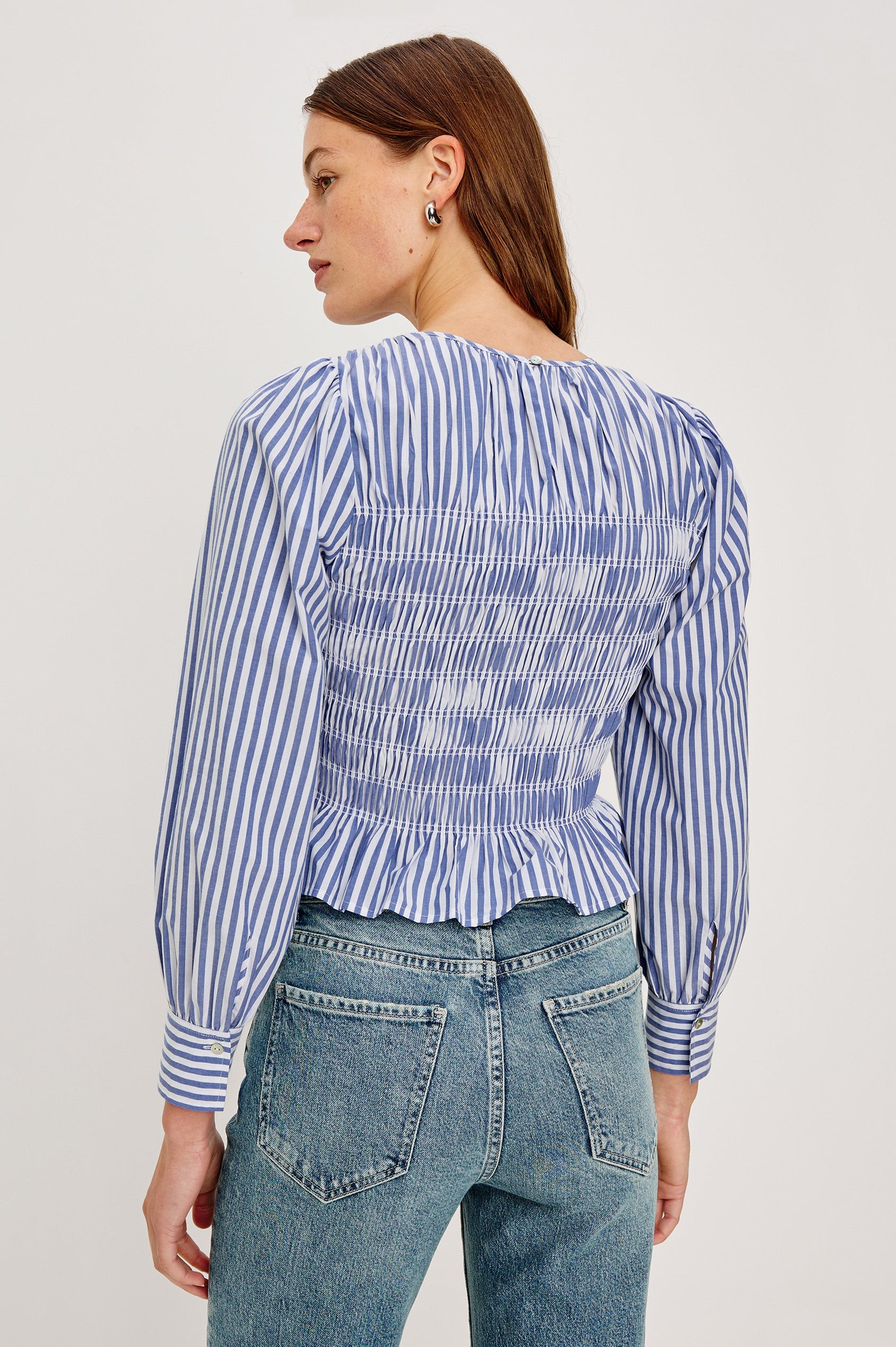 Tabi is a long sleeve crew neck top with puff sleeves and buttons at cuff, the bodice is pleated with a peplum hem in a blue Mariner White Stripe colorway. Back view featured on model.