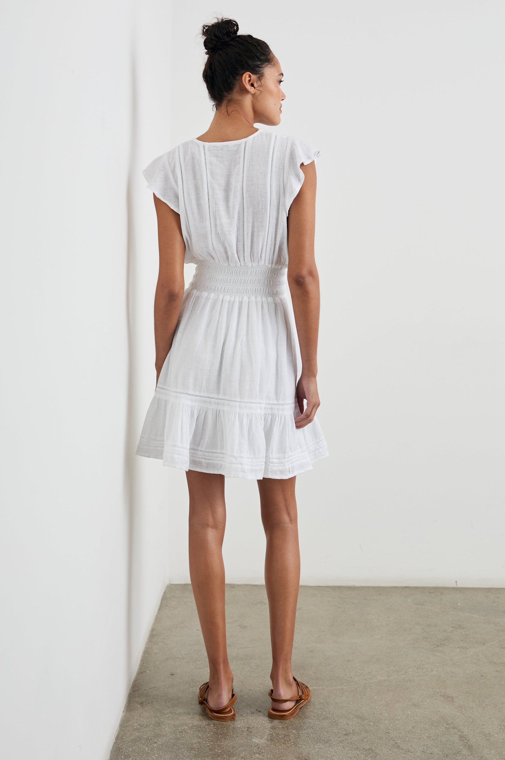 Tara Short Sleeve, Above the Knee Dress with a Smocked Waist, V Neck, Flutter Cap Sleeves, and Tiered Skirt with a Ruffled Hem in White Lace Detail Colorway, with Vertical Lace Inserts on the Bodice and Horizontal Inserts on the Skirt - Back Full Body View Featured on Model 