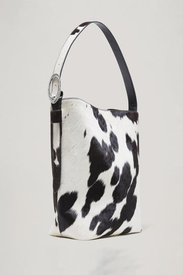 The Torino is a cow-print haircalf leather bucket bag, designed with an adjustable strap and polished silver oval buckle. Front view featured.
