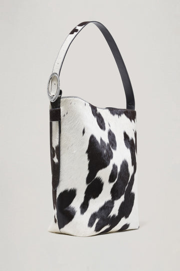 The Torino is a cow-print haircalf leather bucket bag, designed with an adjustable strap and polished silver oval buckle. Front view featured.
