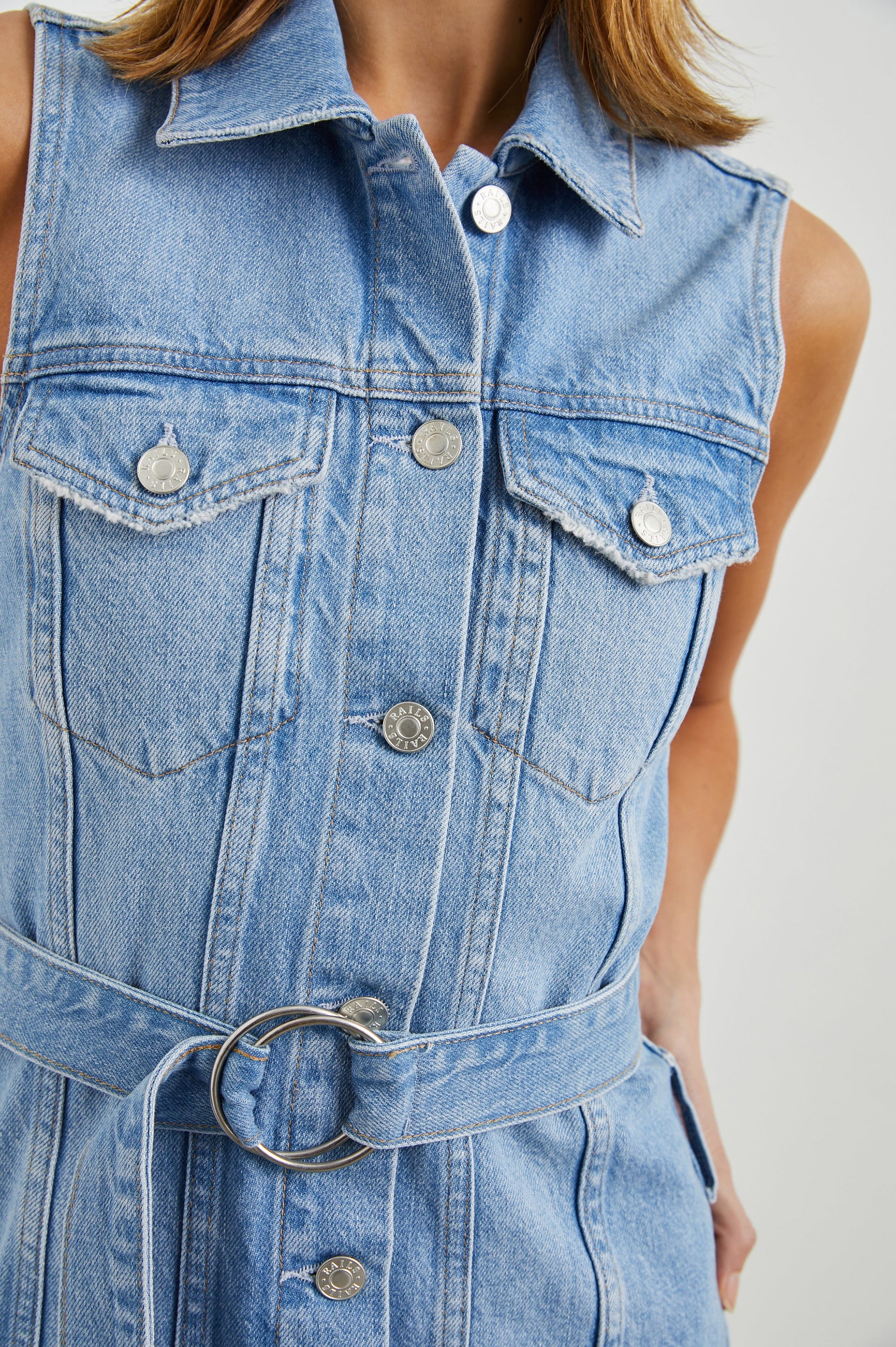 Wilshire Sleeveless Denim Mini Dress with a Full Button Front, Two Chest Button Flap Pockets, a Double Ring Belt, a Collar, and Front Vertical Seams in Blue Denim Colorway - Front View Featured on Model 