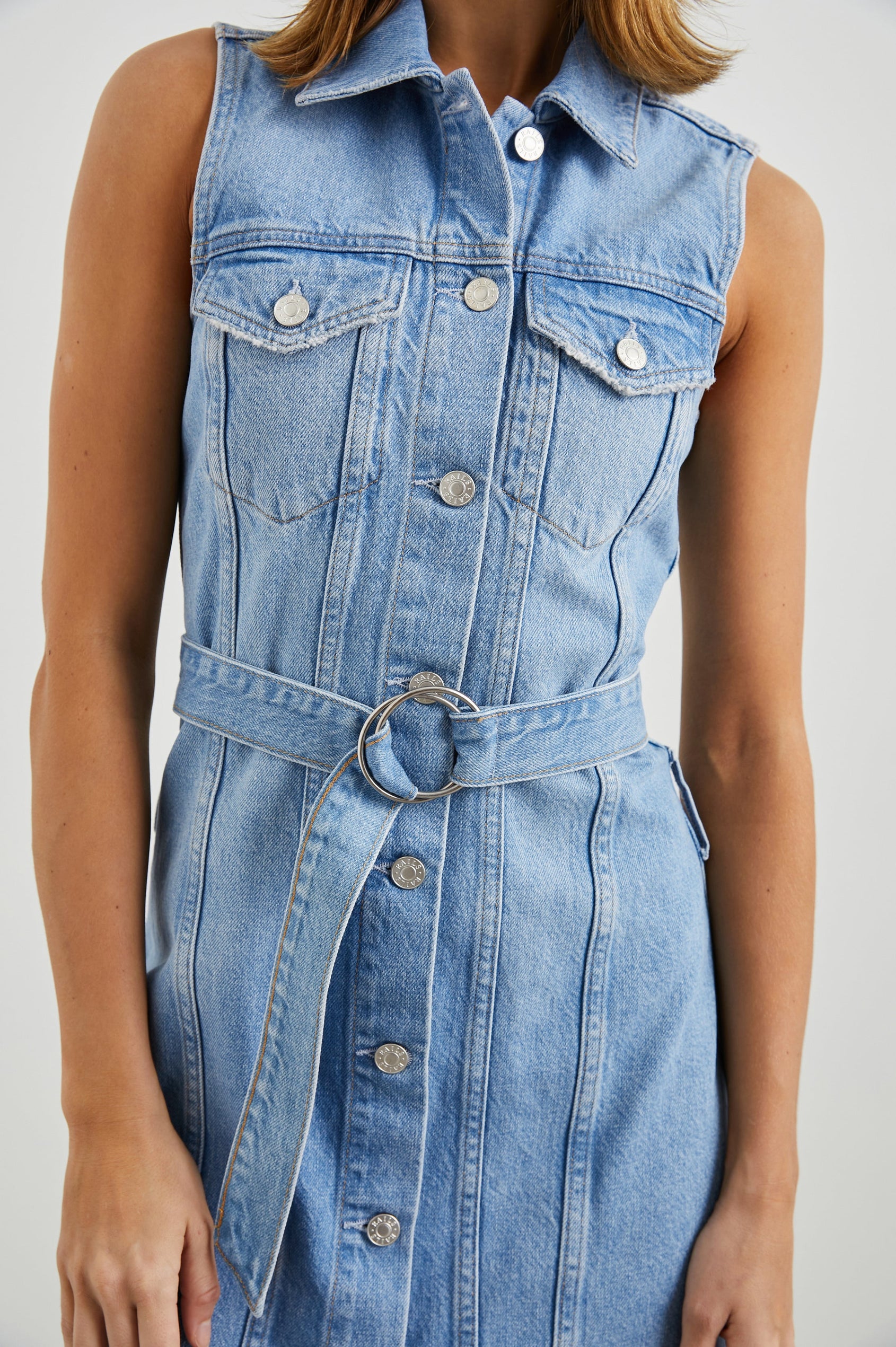 Wilshire Sleeveless Denim Mini Dress with a Full Button Front, Two Chest Button Flap Pockets, a Double Ring Belt, a Collar, and Front Vertical Seams in Blue Denim Colorway - Front View Featured on Model 