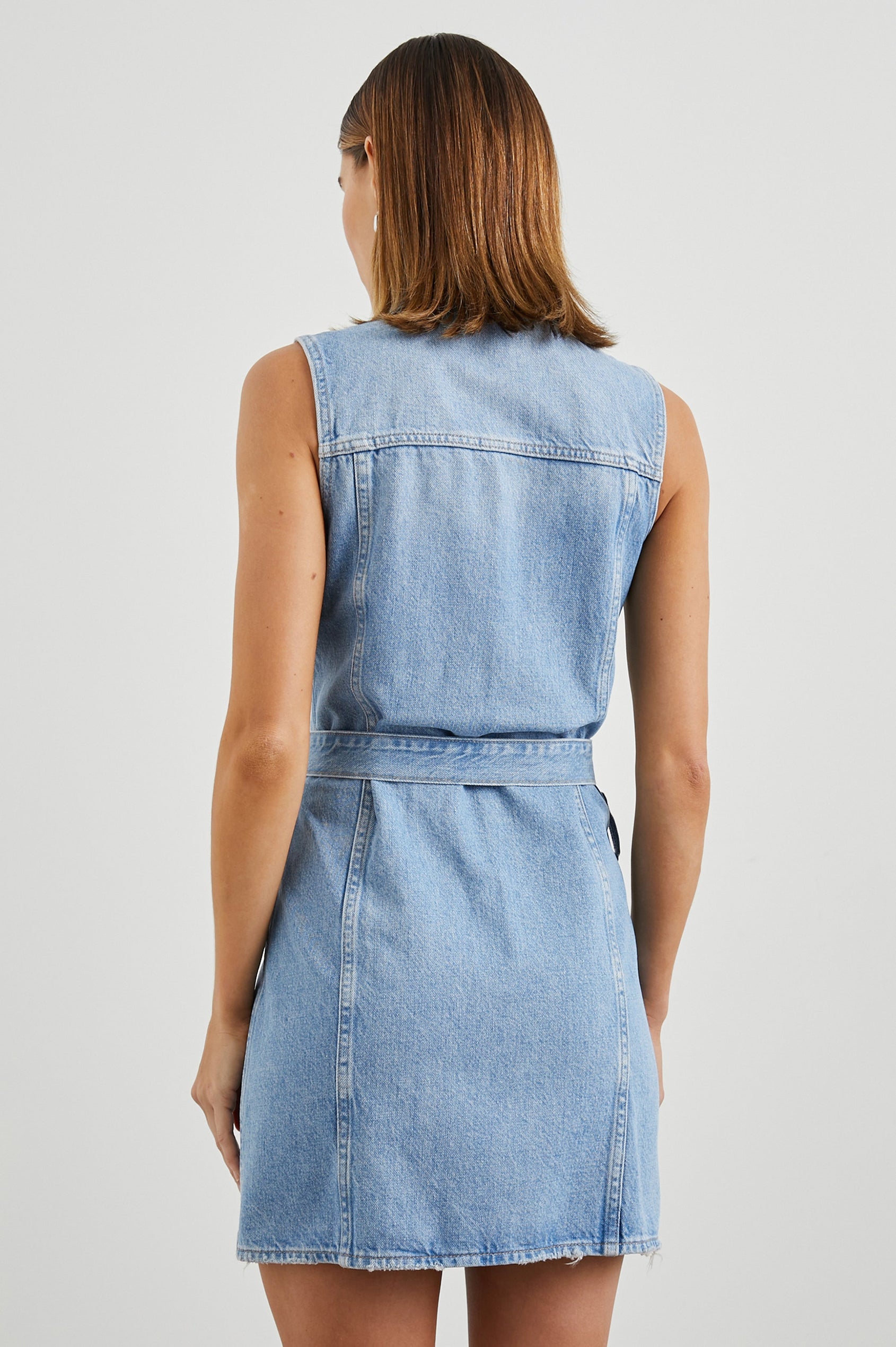 Wilshire Sleeveless Denim Mini Dress with a Full Button Front, Two Chest Button Flap Pockets, a Double Ring Belt, a Collar, and Front Vertical Seams in Blue Denim Colorway - Back View Featured on Model 