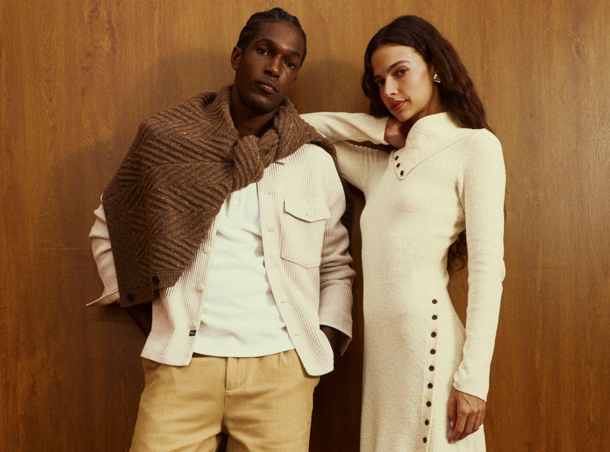 A man and a woman posing together against a warm wood-paneled wall. The man wears a cream ribbed jacket over a white shirt with a brown herringbone knit shawl draped over his shoulders, paired with beige trousers.