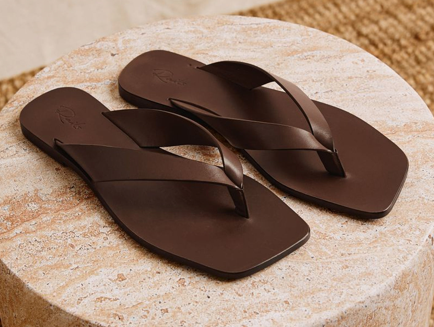 A pair of dark brown leather flip-flops with square toes, displayed on a round beige stone pedestal.
