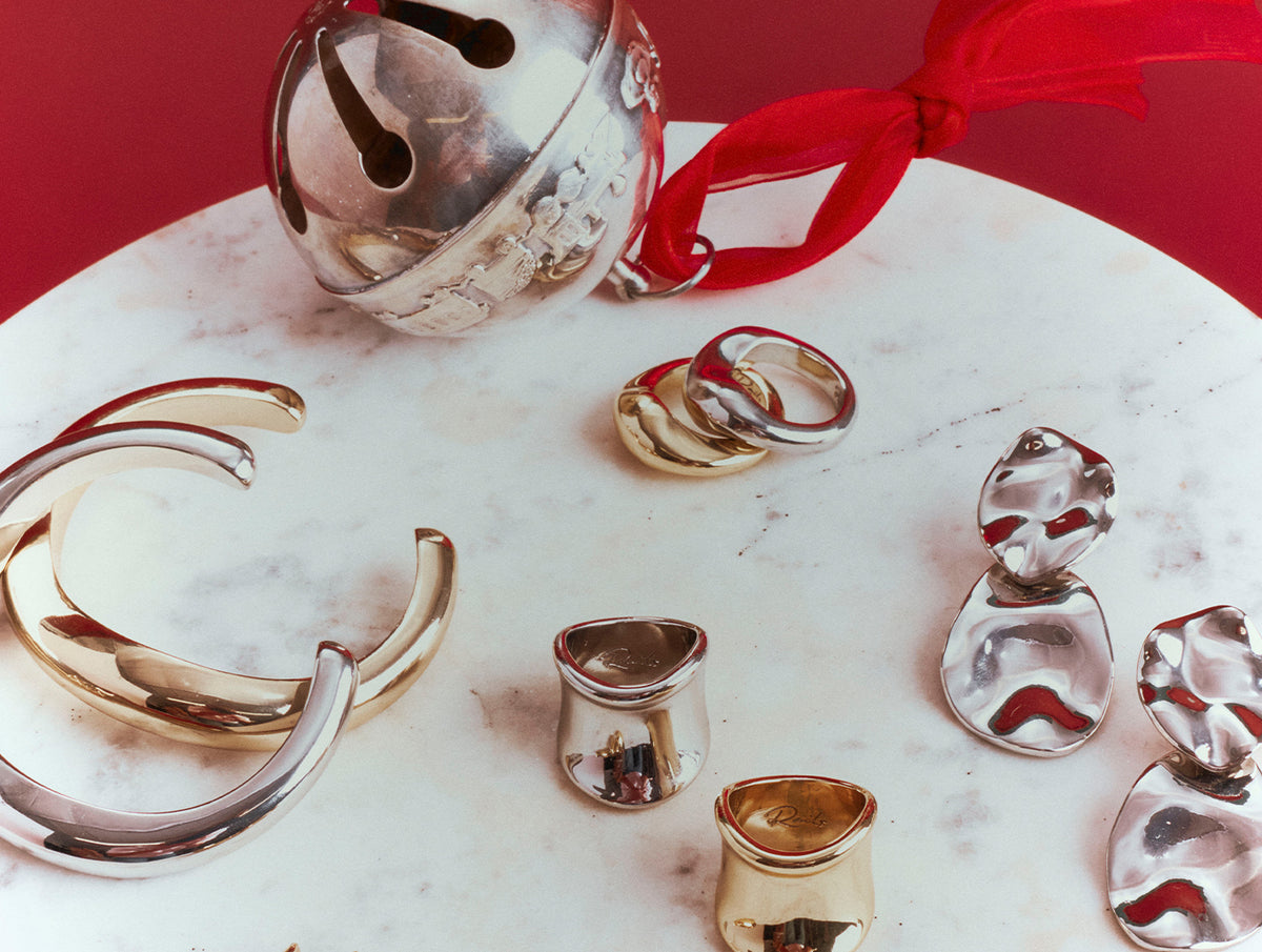 A collection of polished metallic jewelry pieces, including a large silver-toned cuff, a silver bell with a red ribbon, several rings, and small sculptural pieces, arranged on a white marble surface against a solid red background.