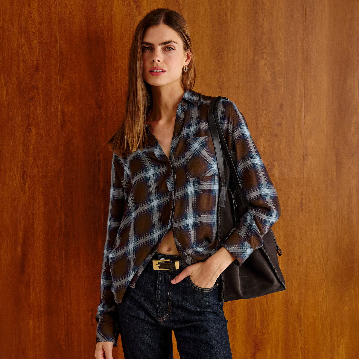A woman stands against a warm wood-paneled wall wearing a brown and blue plaid button-up shirt, dark blue jeans, and a black shoulder bag.