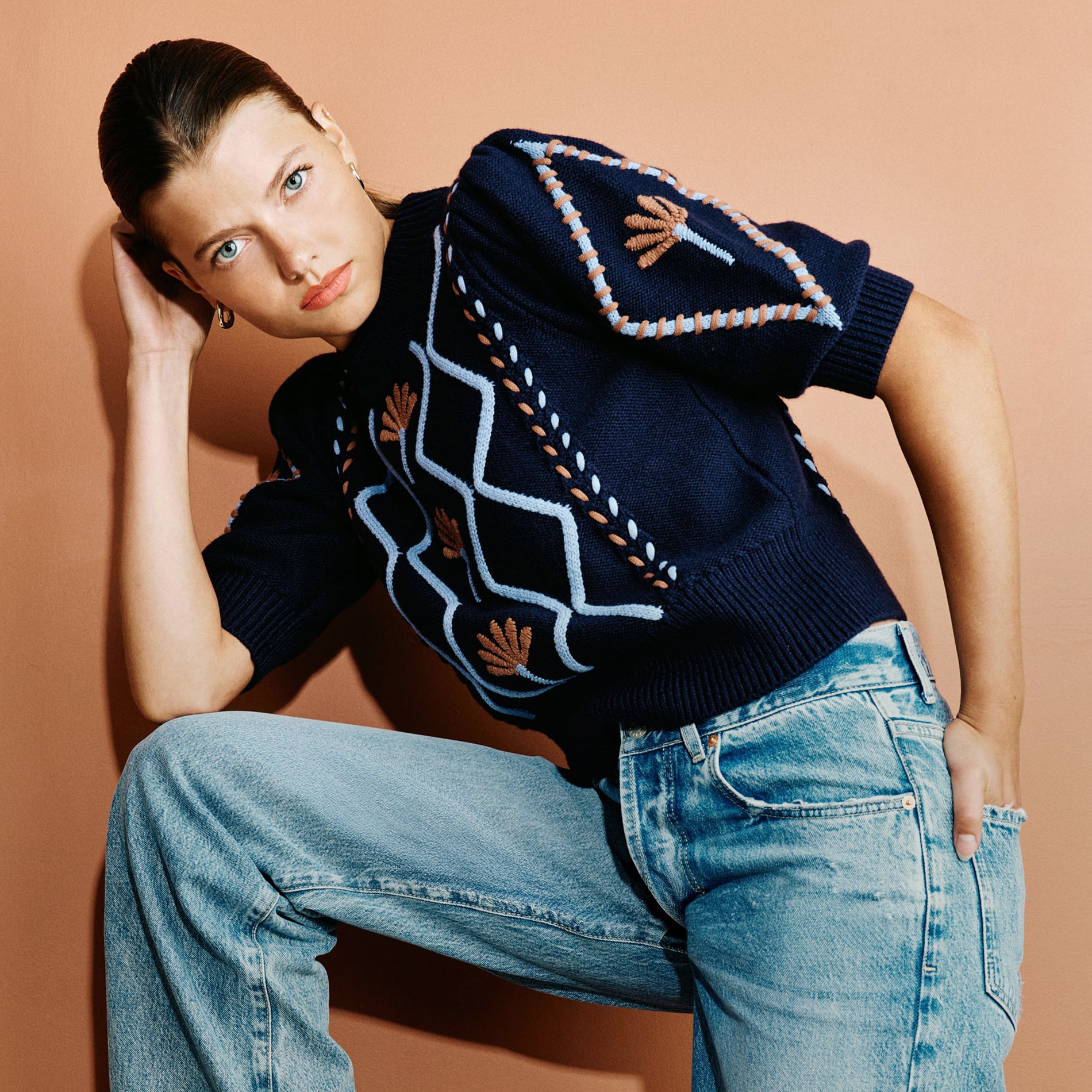 A woman poses in a navy blue short-sleeve knit sweater with geometric and floral patterns, paired with light blue jeans, set against a solid peach background.