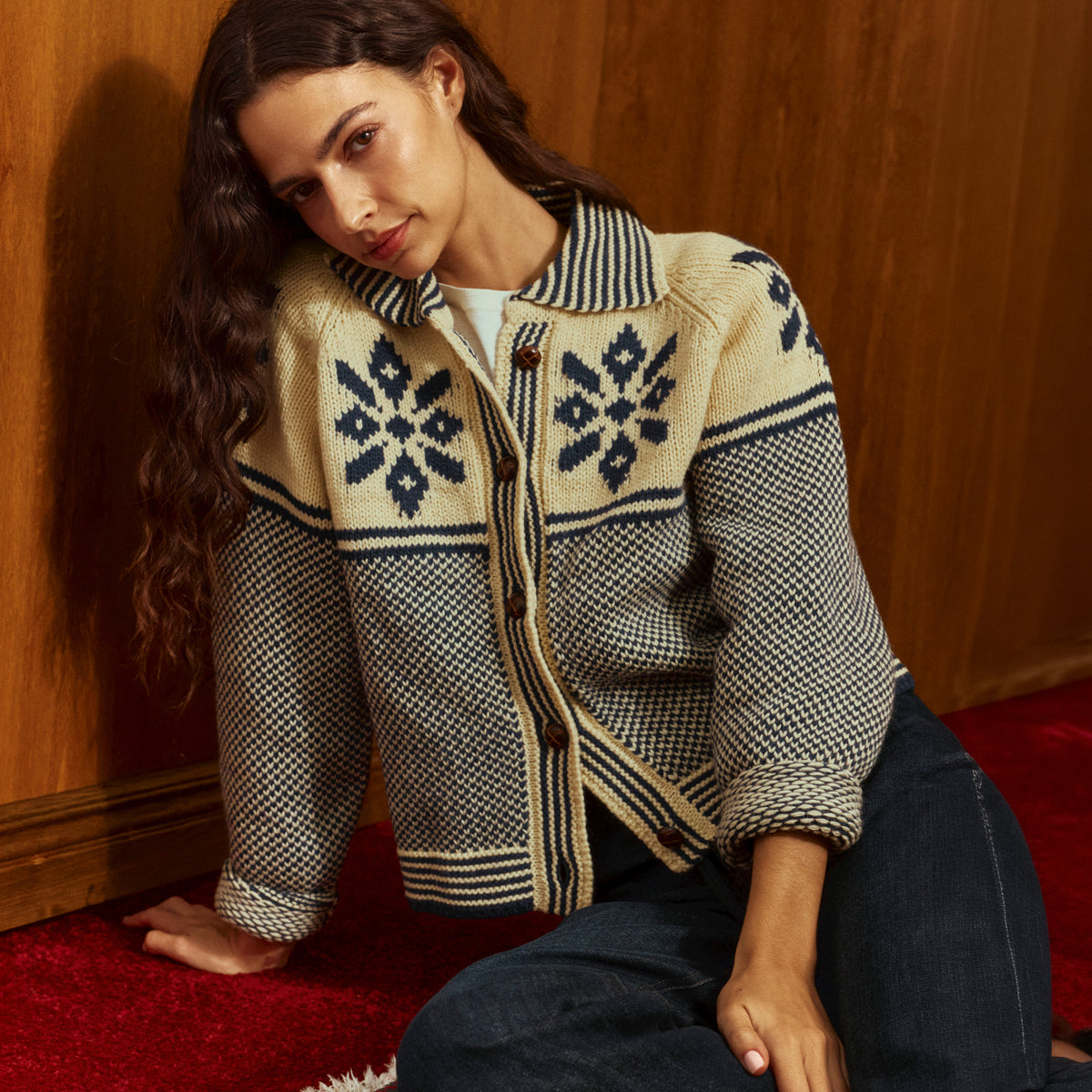 A woman kneels on a red carpet against a wooden wall, wearing a cream and navy patterned knit cardigan with dark blue jeans.