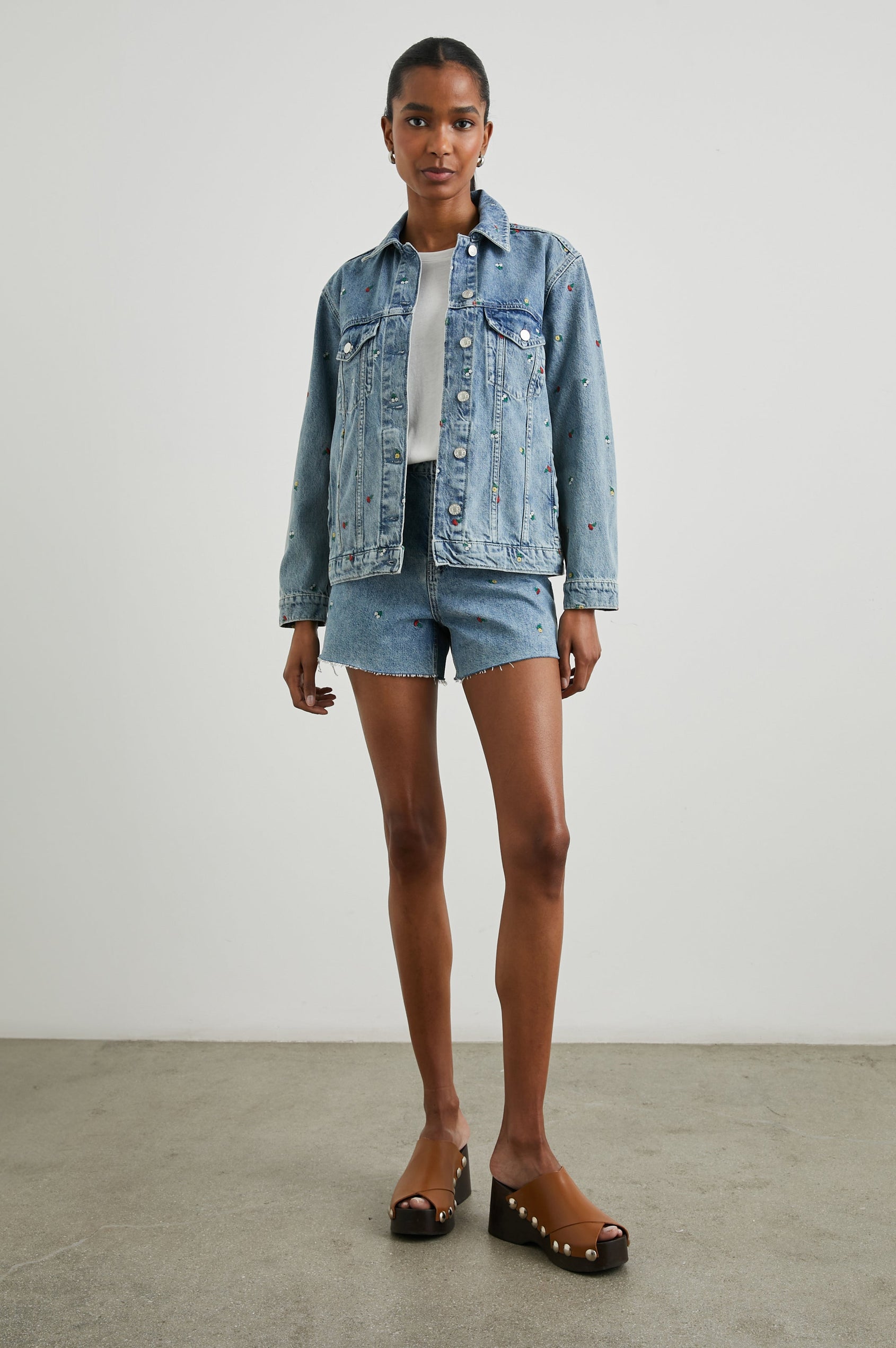 Zuma Denim Shorts with a Raw Hem, Belt Loops, Two Front Pockets Plus a Coin Pocket and Two Back Patch Pockets in Flower Field Colorway, with Embroidered Yellow, White and Red Daisies - Front Full Body View Featured on Model 