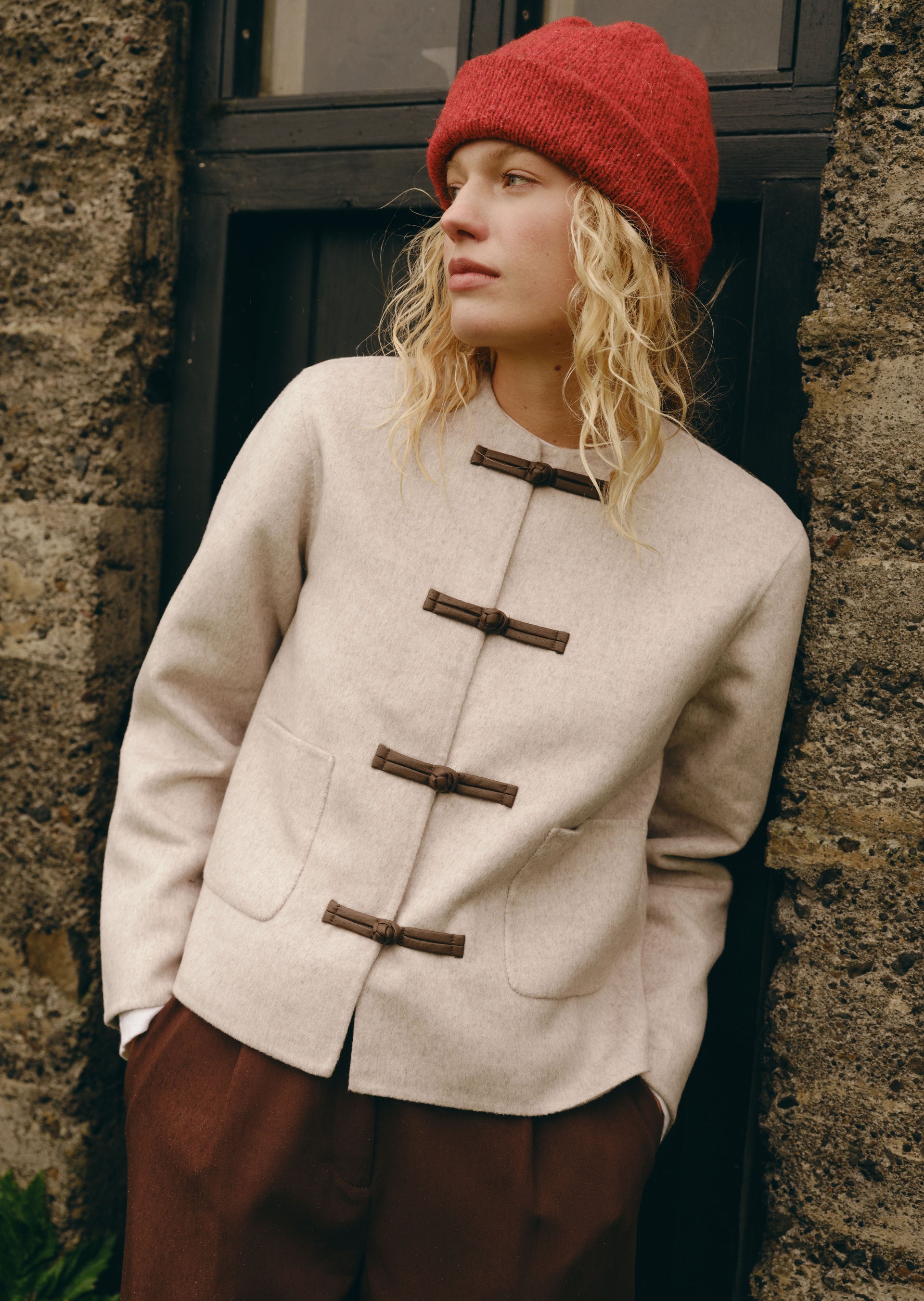 A woman wearing a cream-colored wool jacket with dark brown toggle closures, a red knit beanie, and brown trousers, leaning against a rustic stone wall.