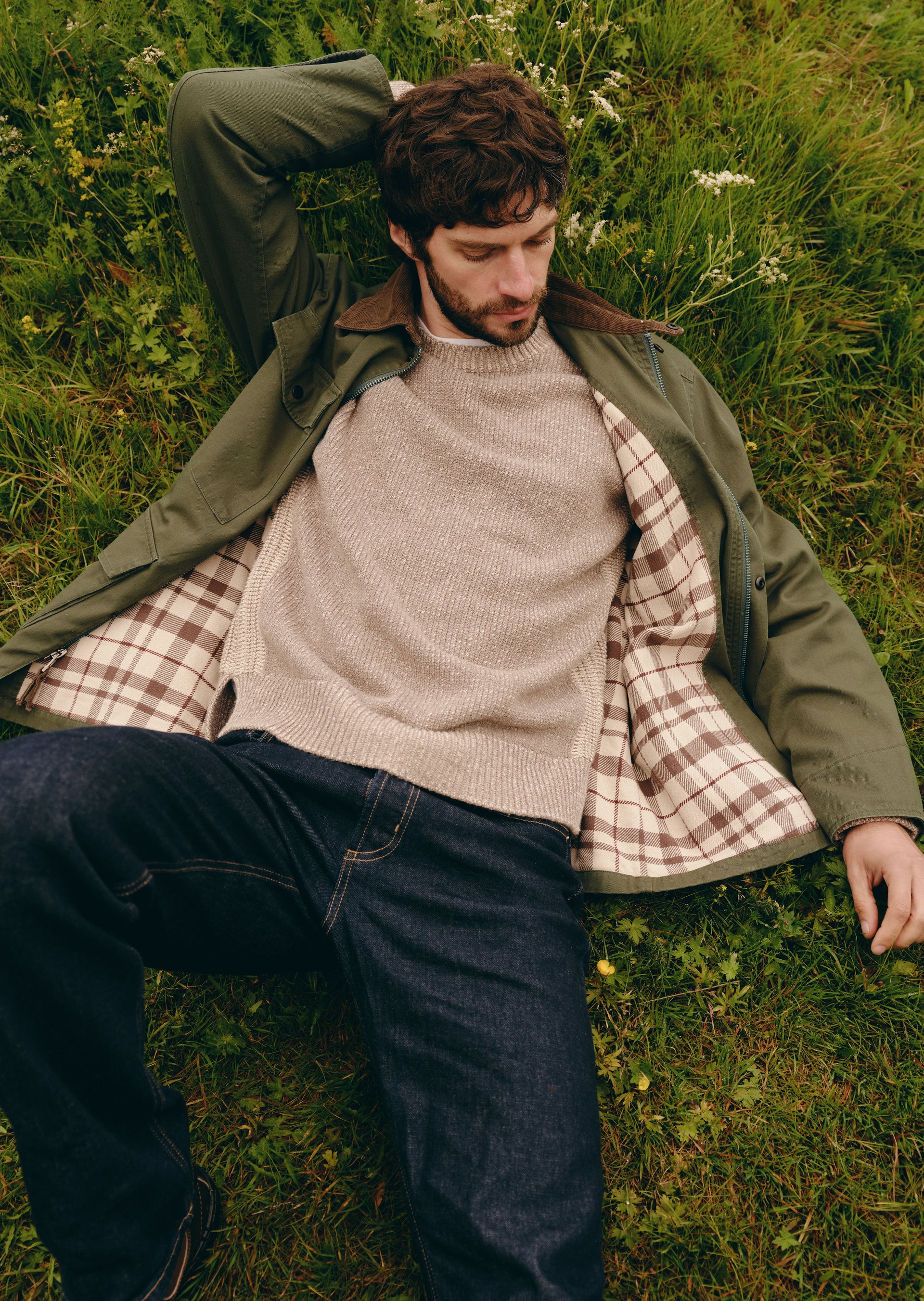 A man lying on green grass wearing an olive green jacket over a beige knit sweater and a brown-and-white plaid shirt, paired with dark blue jeans.