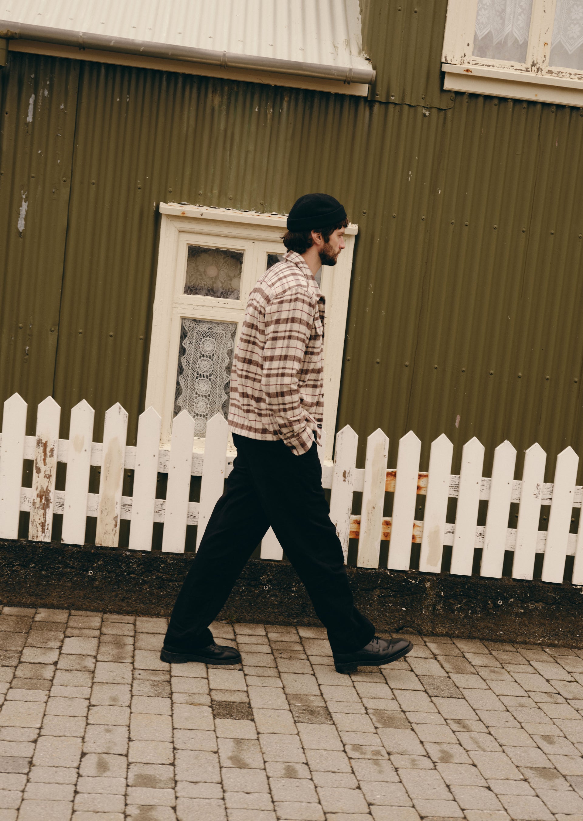A man wearing a beige and brown plaid flannel shirt, black pants, and a black beanie walks past a white picket fence in front of a dark green corrugated metal building.