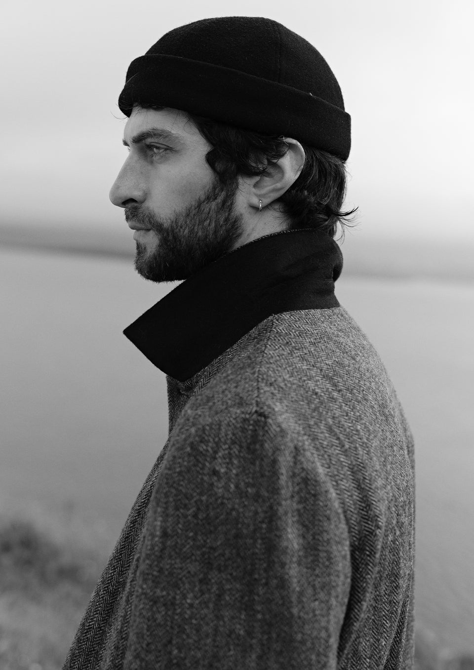 A man in profile wearing a black knit beanie, a dark high-neck collar, and a textured gray coat, photographed outdoors against a soft, overcast background.