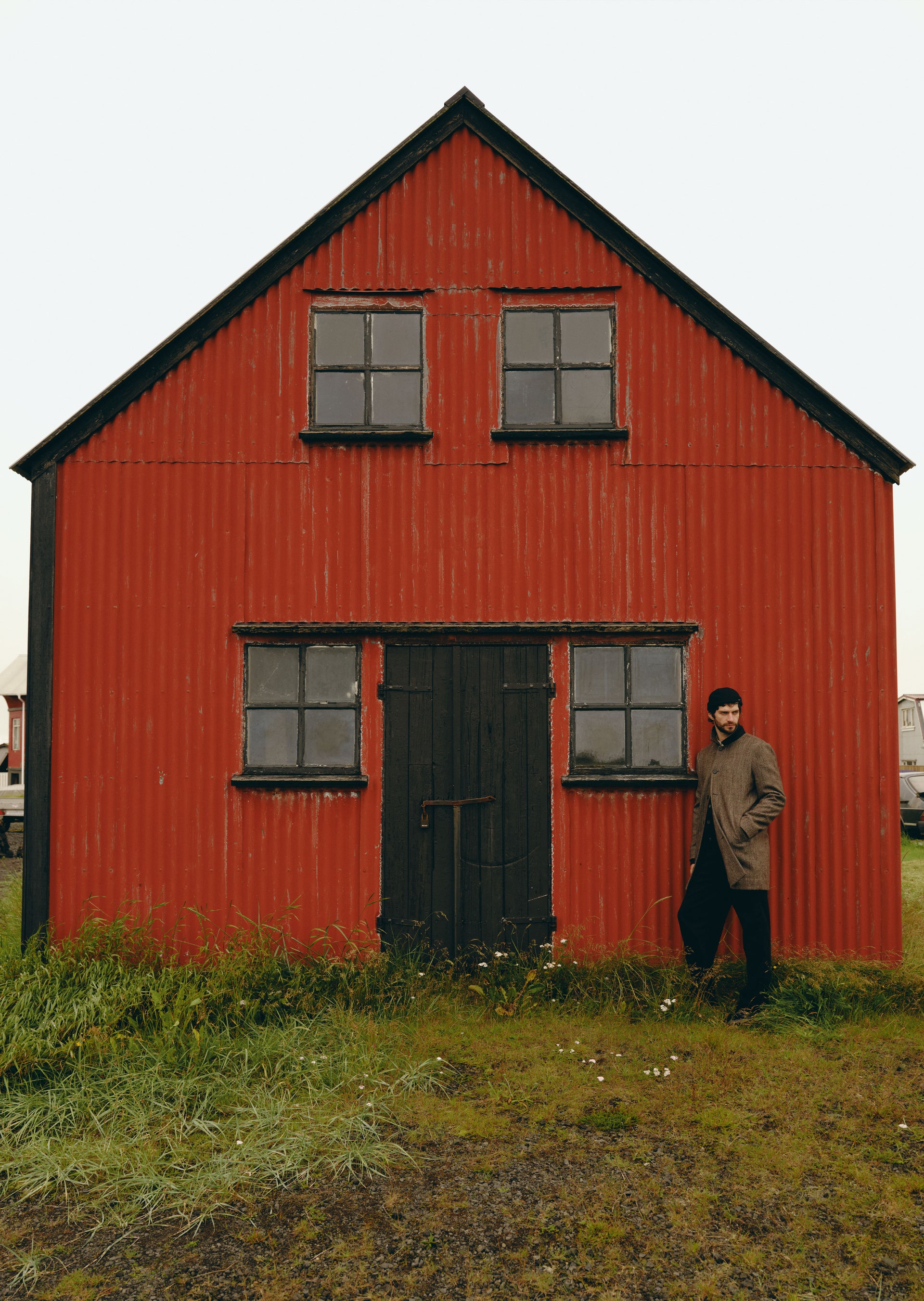 A man in a brown jacket and black beanie stands next to a red wooden house with a dark door and windows, set against an overcast sky.