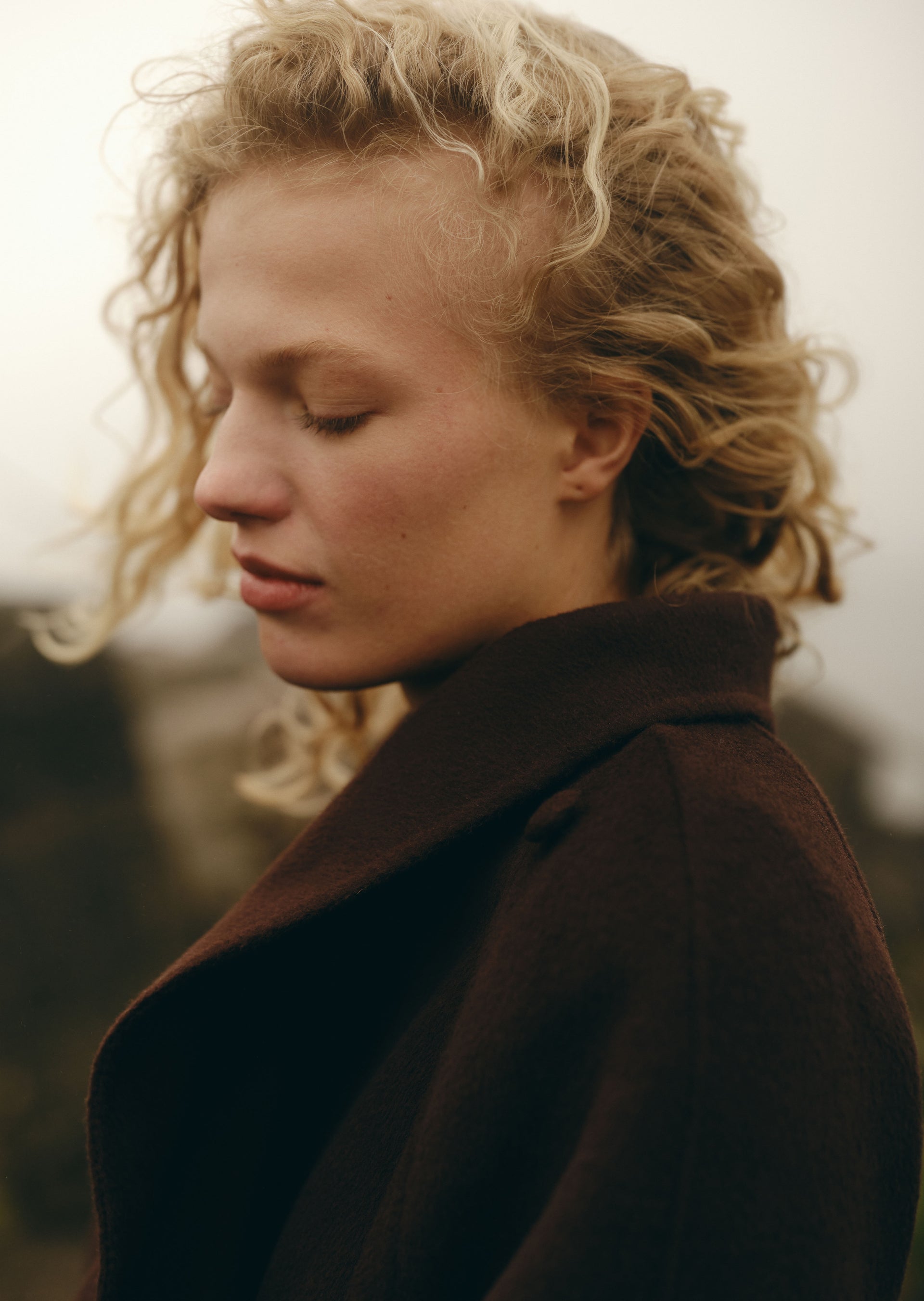 A close-up portrait of a woman with curly blonde hair wearing a dark brown coat, shown in profile against a soft, muted outdoor background.