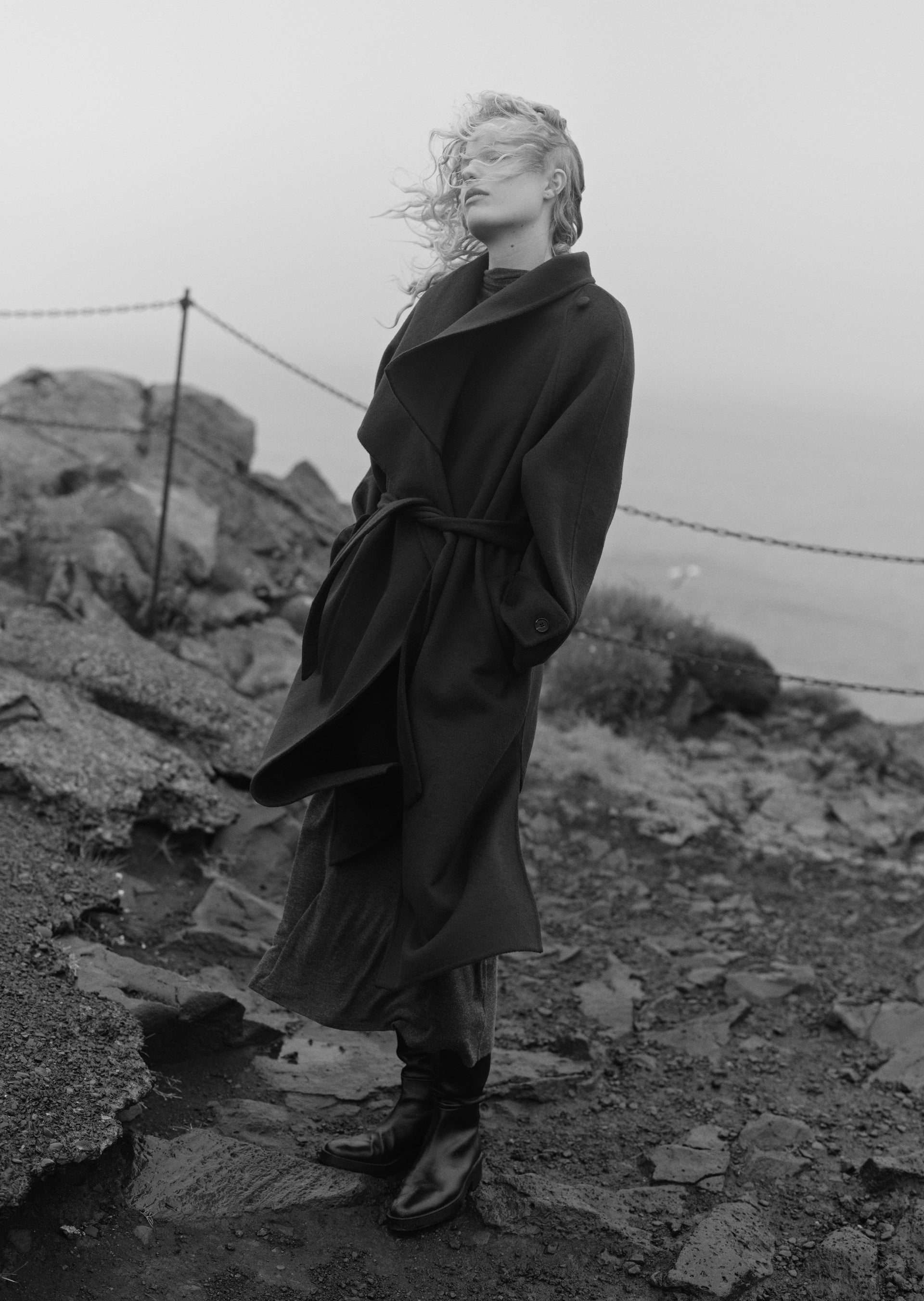 A black and white editorial photograph of a model wearing a long dark coat and boots, standing on a rocky, foggy outdoor terrain.