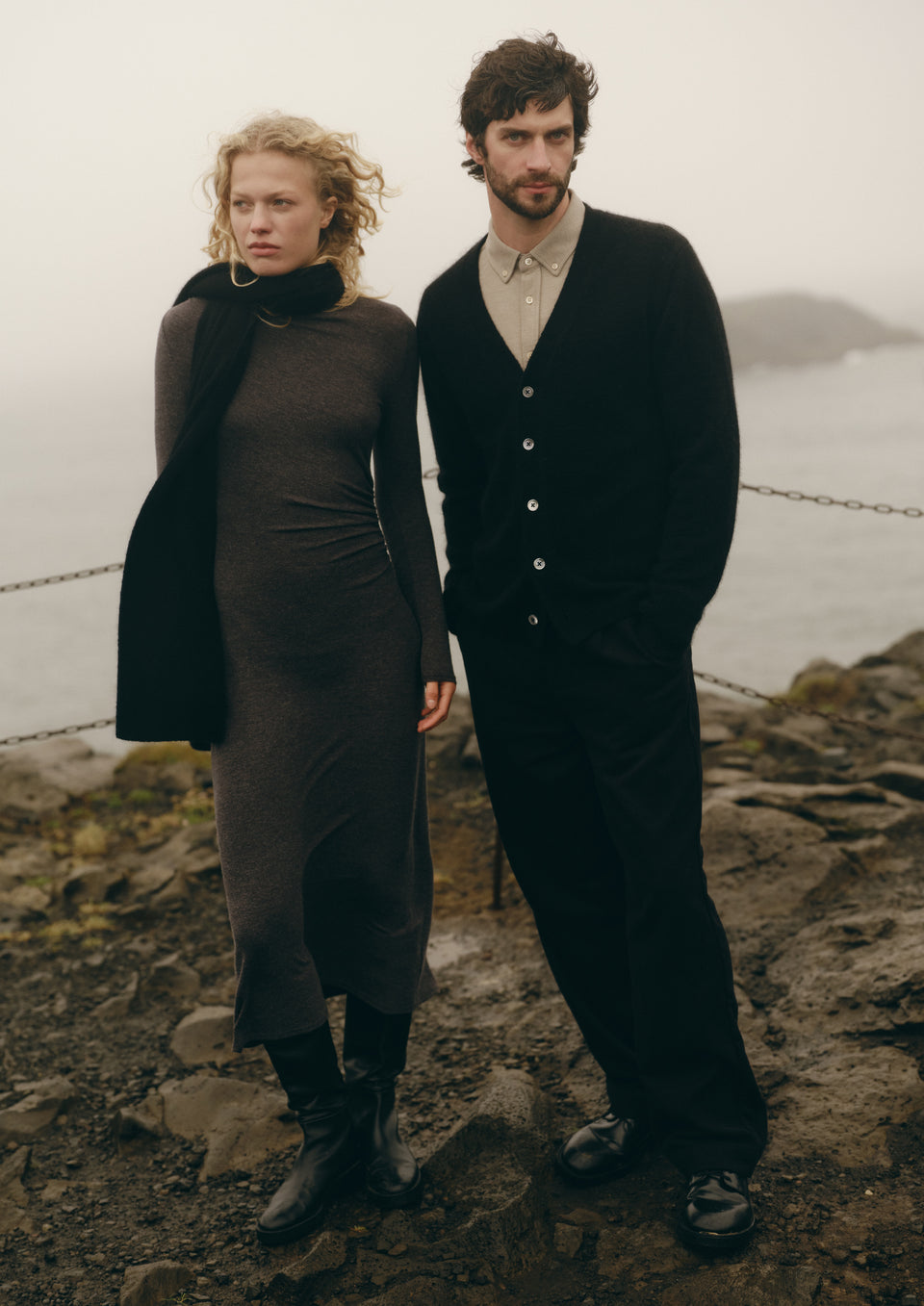A man and a woman standing together outdoors on a rocky shoreline with foggy water in the background. The woman wears a dark long-sleeve dress with a black scarf and black boots, and the man wears a black cardigan over a light shirt with black trousers.