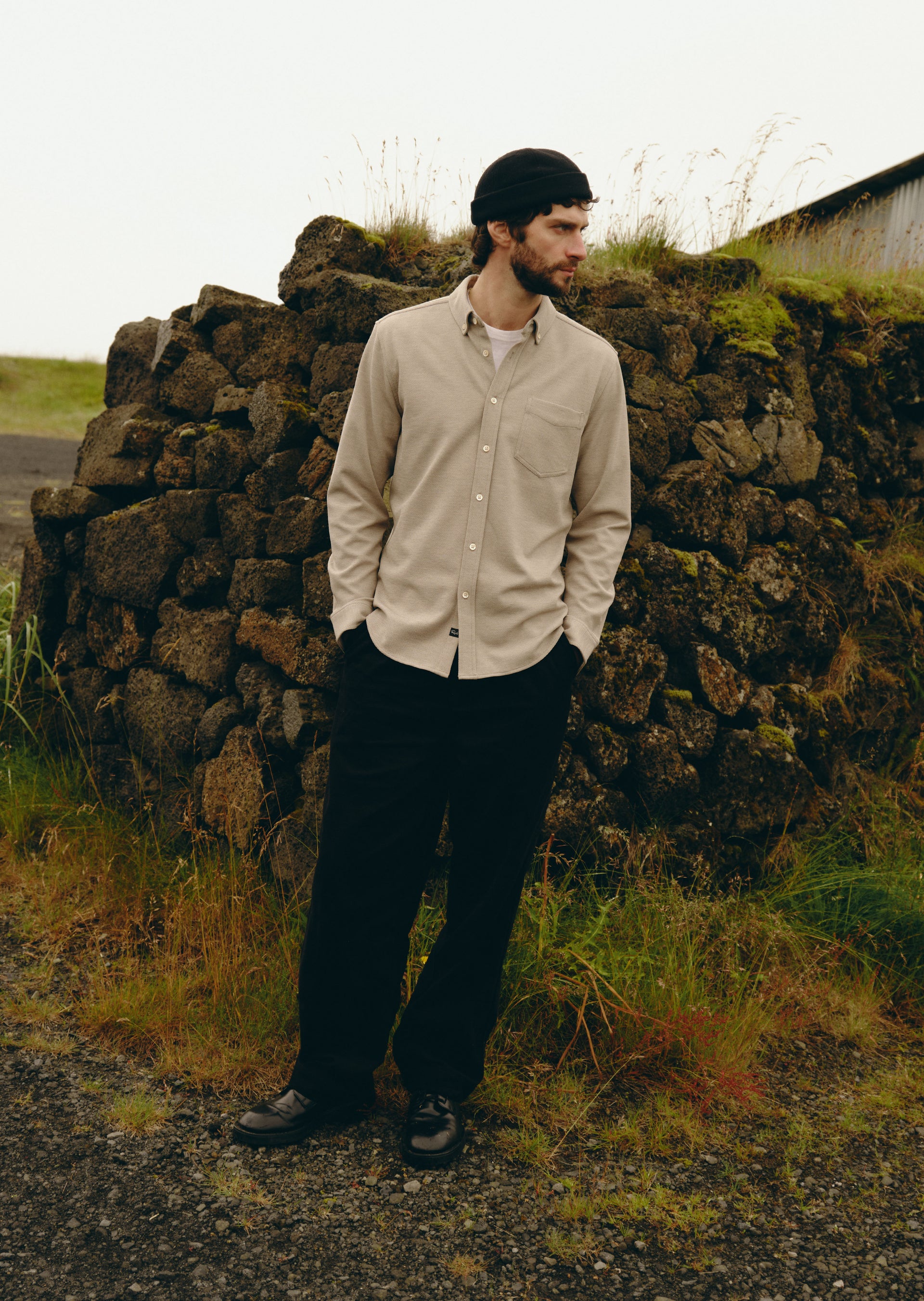 A man wearing a light beige long-sleeve button-down shirt, black pants, and a black beanie stands outdoors in front of a rustic stone wall.