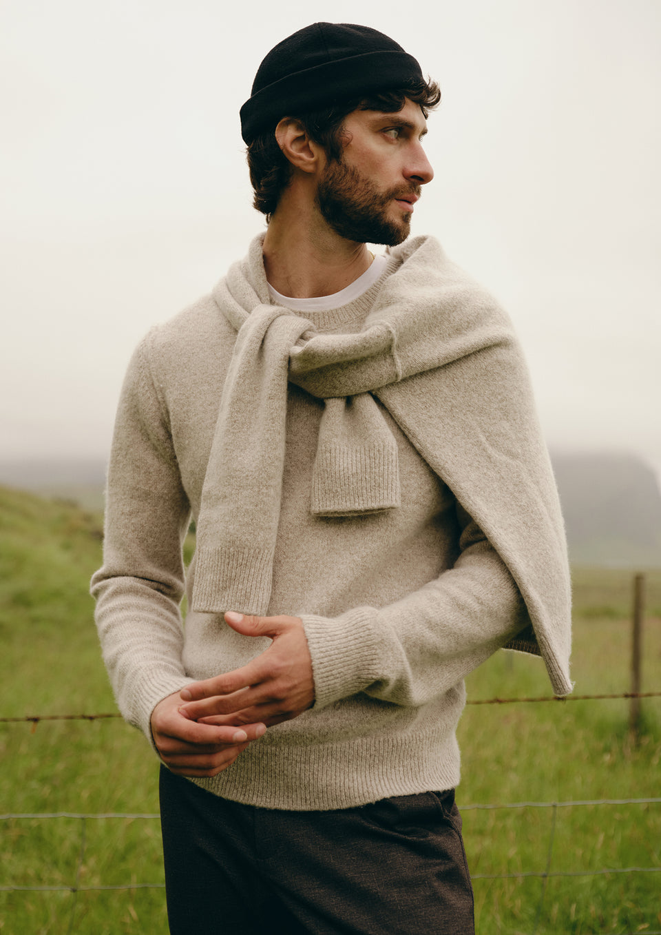 A man wearing a light beige knit sweater and matching draped scarf, standing outdoors in a grassy field under an overcast sky.