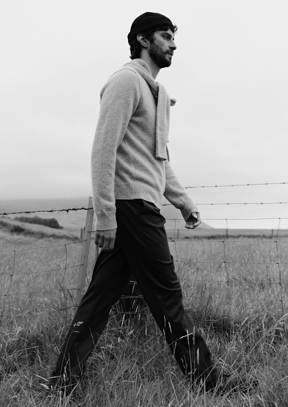 A man wearing a light beige knit sweater and matching draped scarf, standing outdoors in a grassy field under an overcast sky.