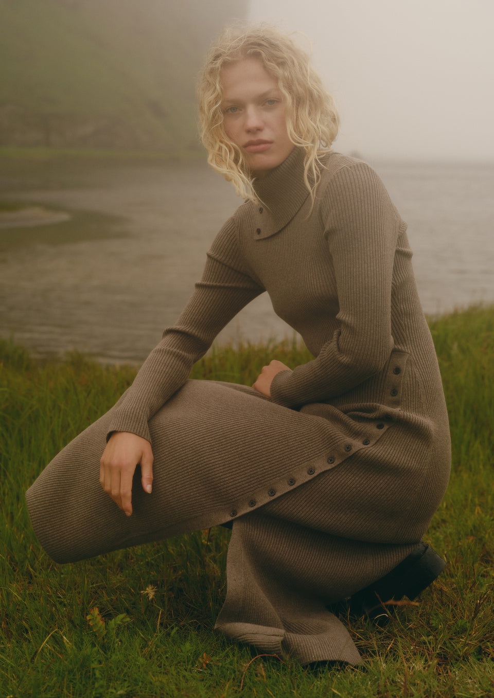 A woman crouches outdoors on grass near water, wearing a ribbed long-sleeve turtleneck dress in a taupe-brown color with decorative buttons along the side.
