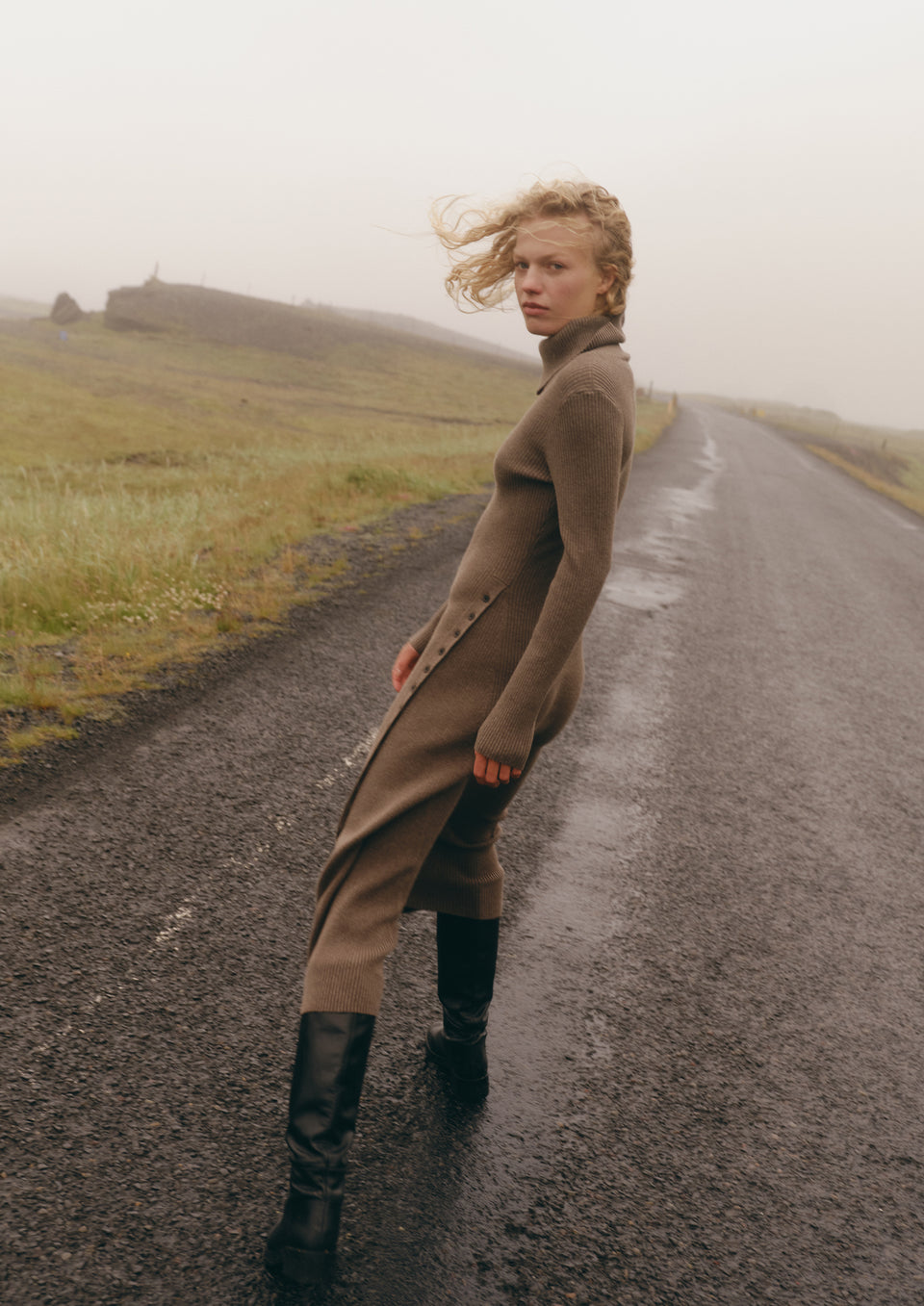 A woman stands on a wet country road wearing a long taupe-brown turtleneck dress with black knee-high boots in a misty outdoor setting.
