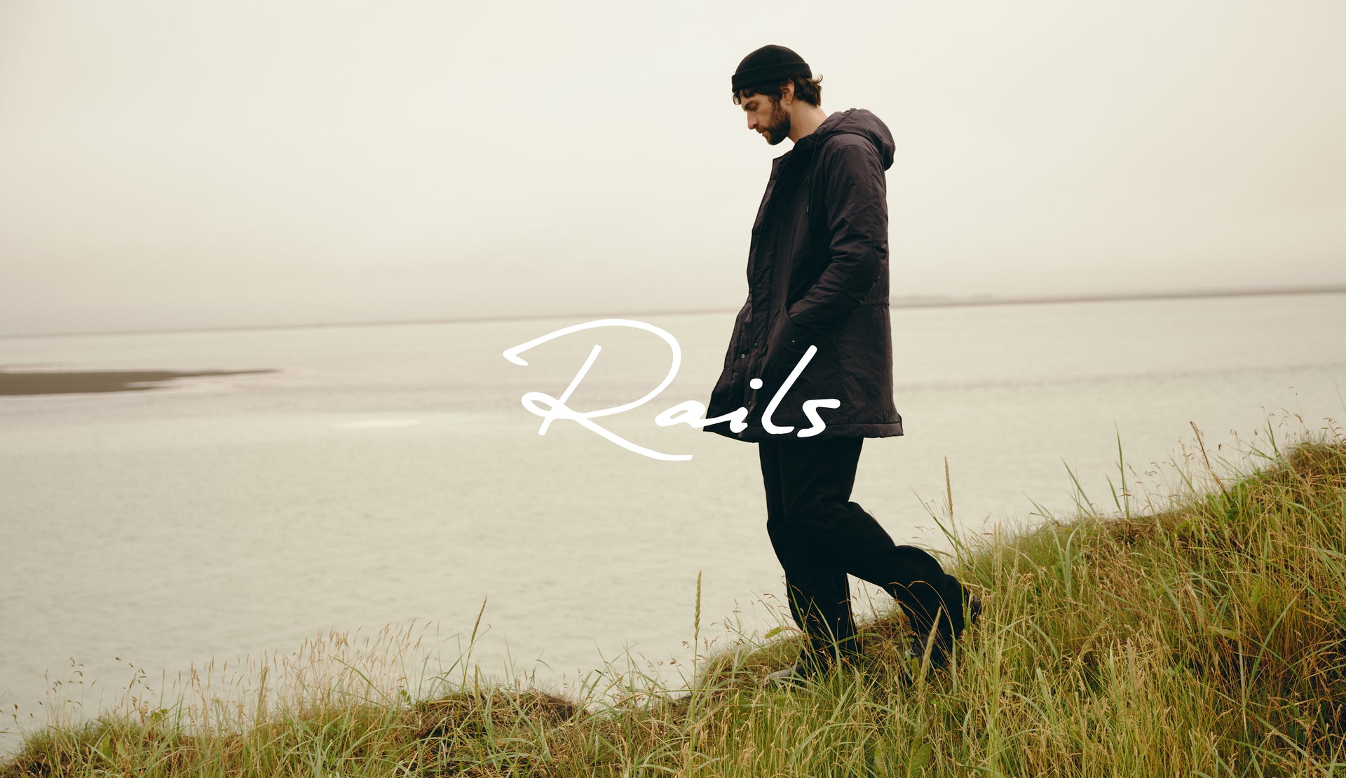 A man wearing a dark hooded jacket and black pants walks along a grassy shoreline next to a calm body of water under an overcast sky. A white script logo reading 'Rails' is overlaid across the center.