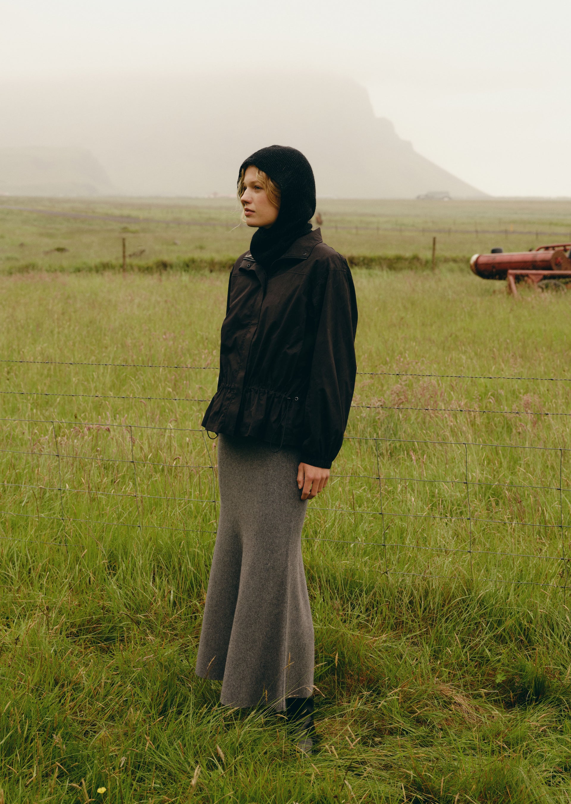 A model stands in a grassy field wearing a black hooded jacket and a long gray skirt, with a misty mountain in the background.