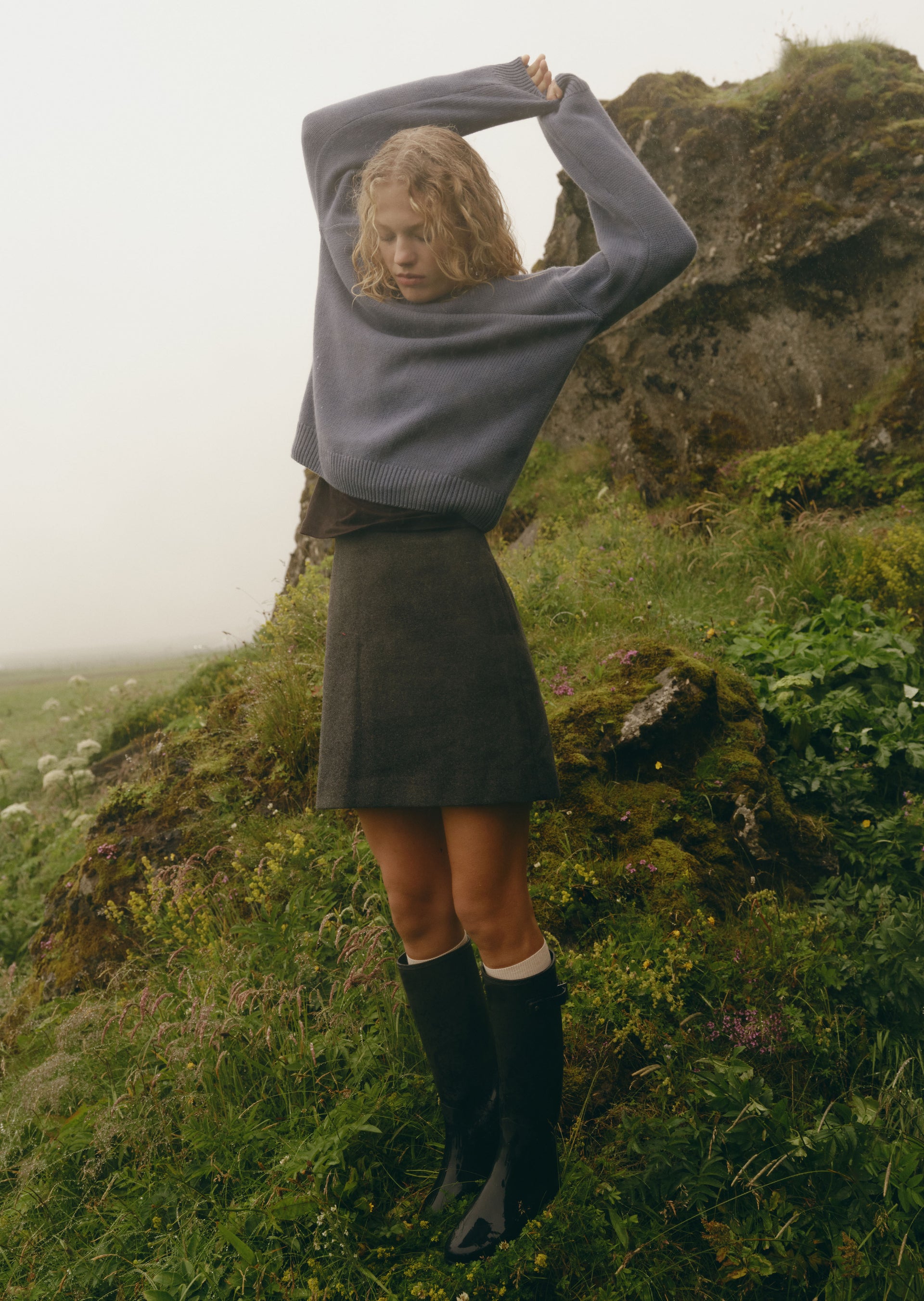 A woman stands outdoors on a grassy, mossy hill wearing a loose gray knit sweater, a dark gray mini skirt, and tall black rain boots.