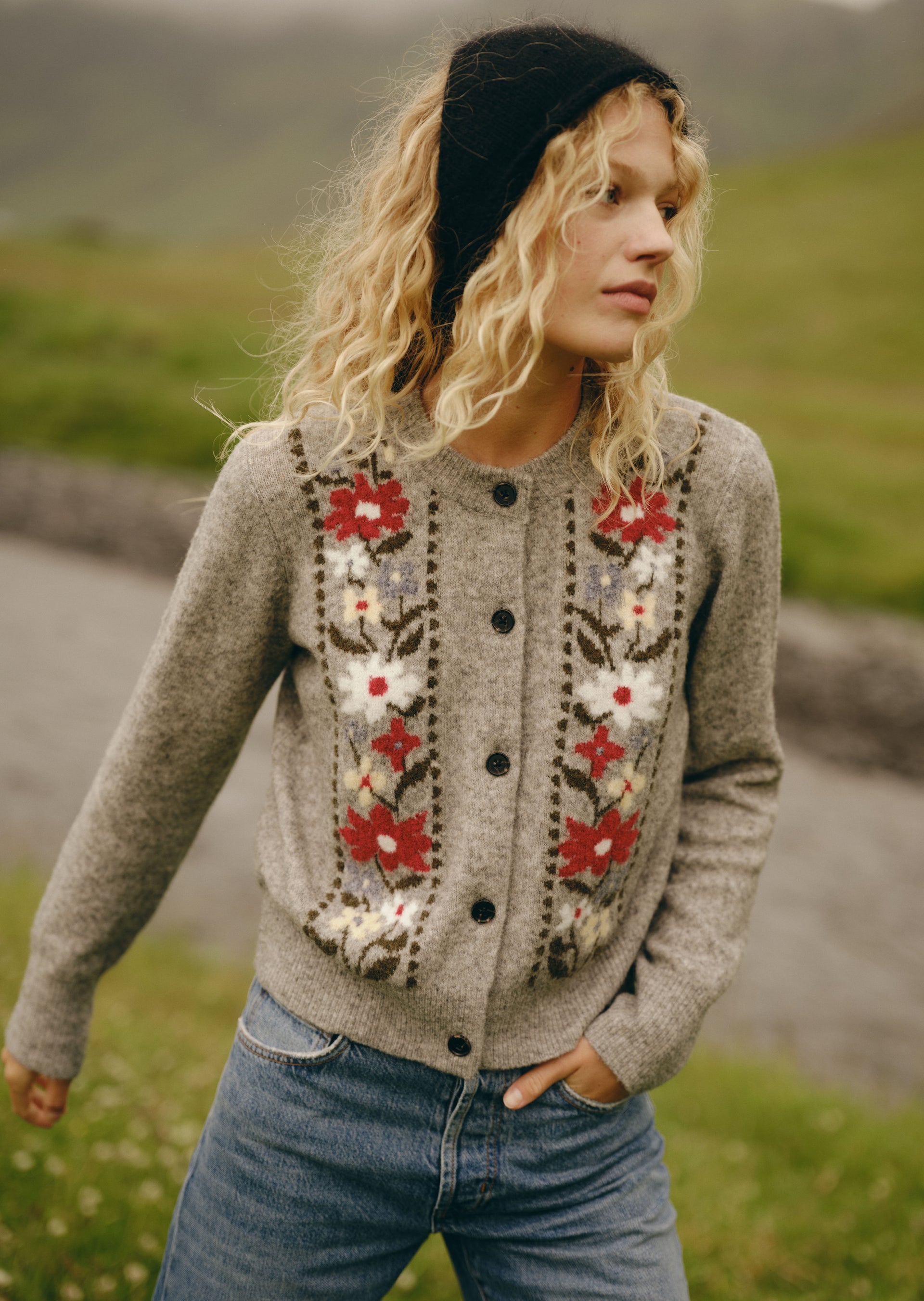 A woman wearing a gray knit button-front cardigan with red, white, and beige floral embroidery, blue jeans, and a black beanie, photographed outdoors in a blurred grassy landscape.