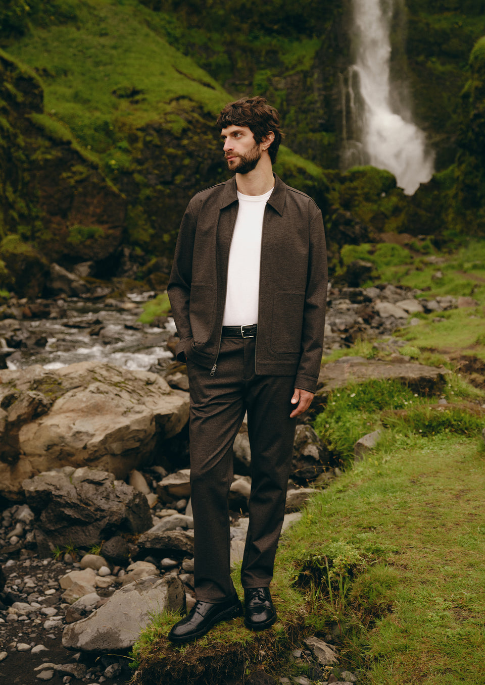 A man wearing a dark brown leather jacket, matching brown trousers, a white t-shirt, and black shoes stands on a rocky path with a waterfall and green forest in the background.