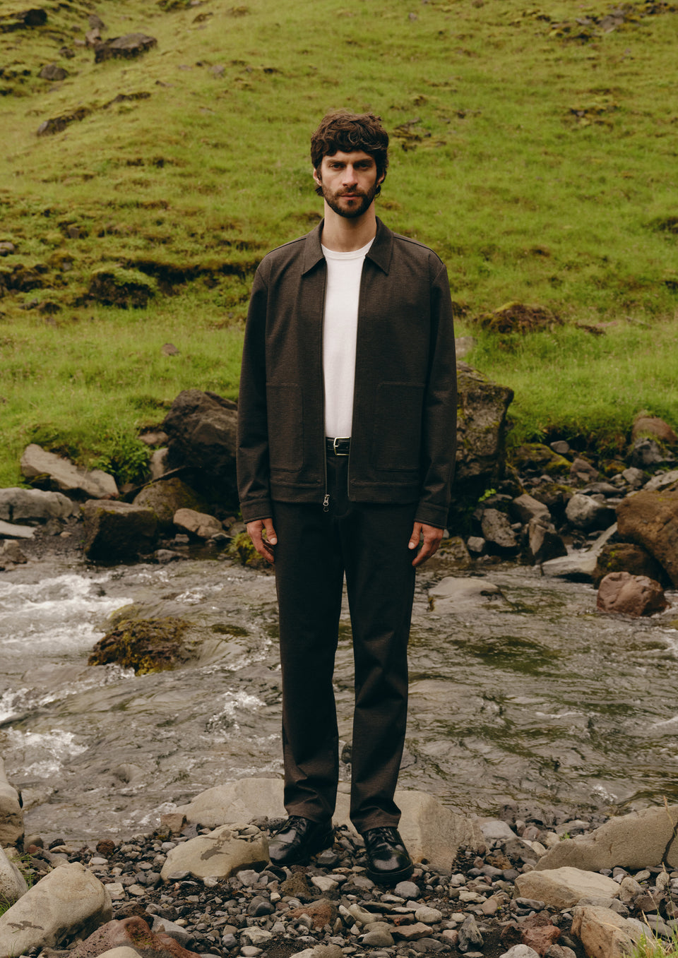 A man stands on a rocky stream in a grassy outdoor setting, wearing a dark brown jacket over a white t-shirt, dark brown trousers, and black shoes.