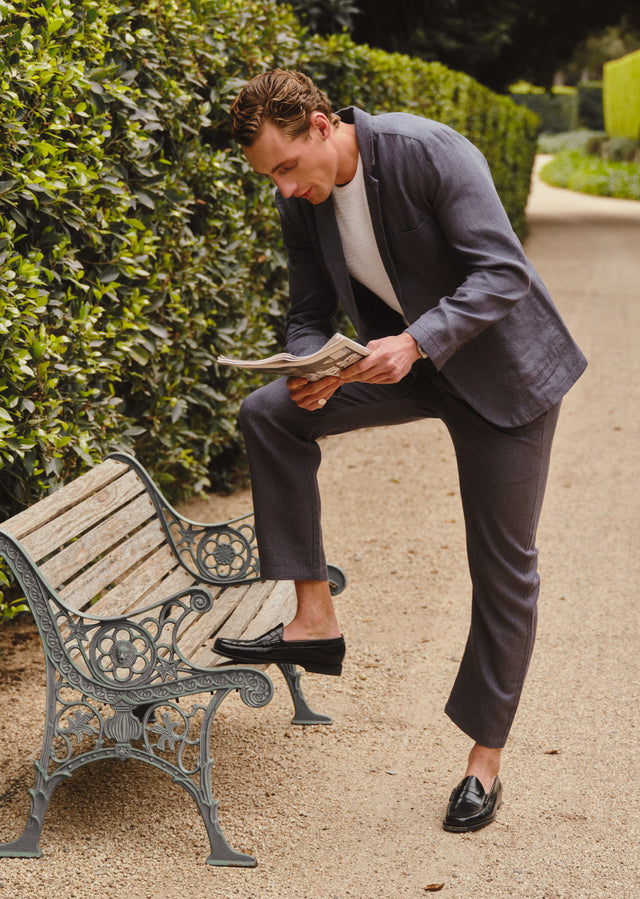 Man in a blue suit sitting on a bench reading a newspaper with greenery in the background