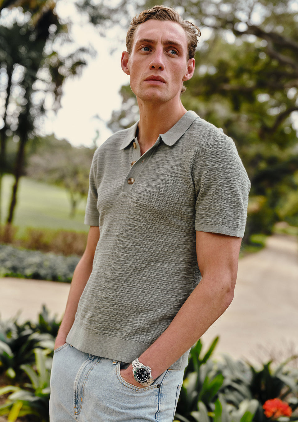 Man wearing a green polo shirt and light blue jeans standing outdoors with greenery in the background