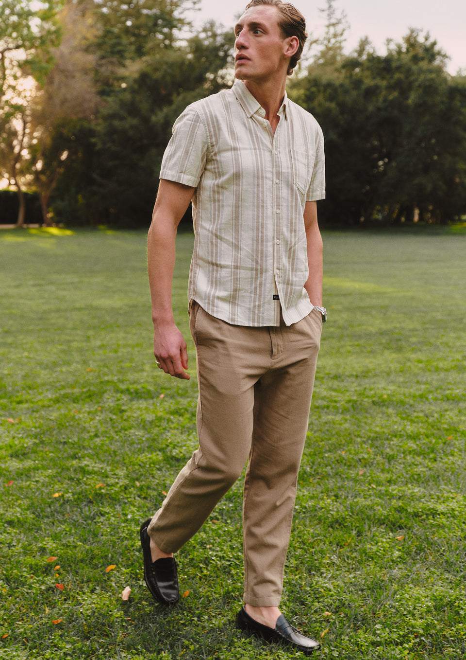 Man wearing a light-colored shirt and beige pants standing on grass with trees in the background