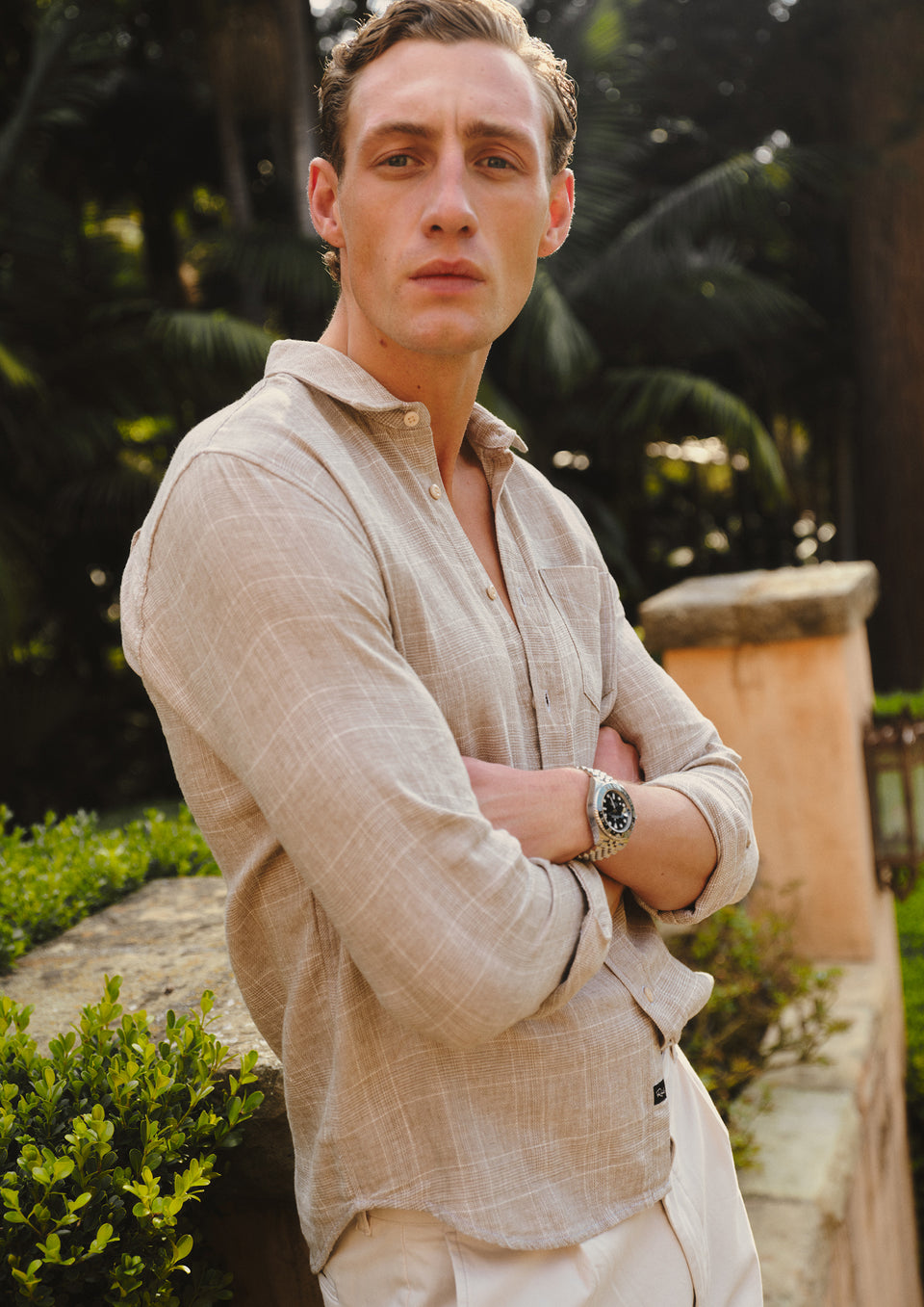 Man wearing a beige shirt and white pants standing outdoors with greenery in the background