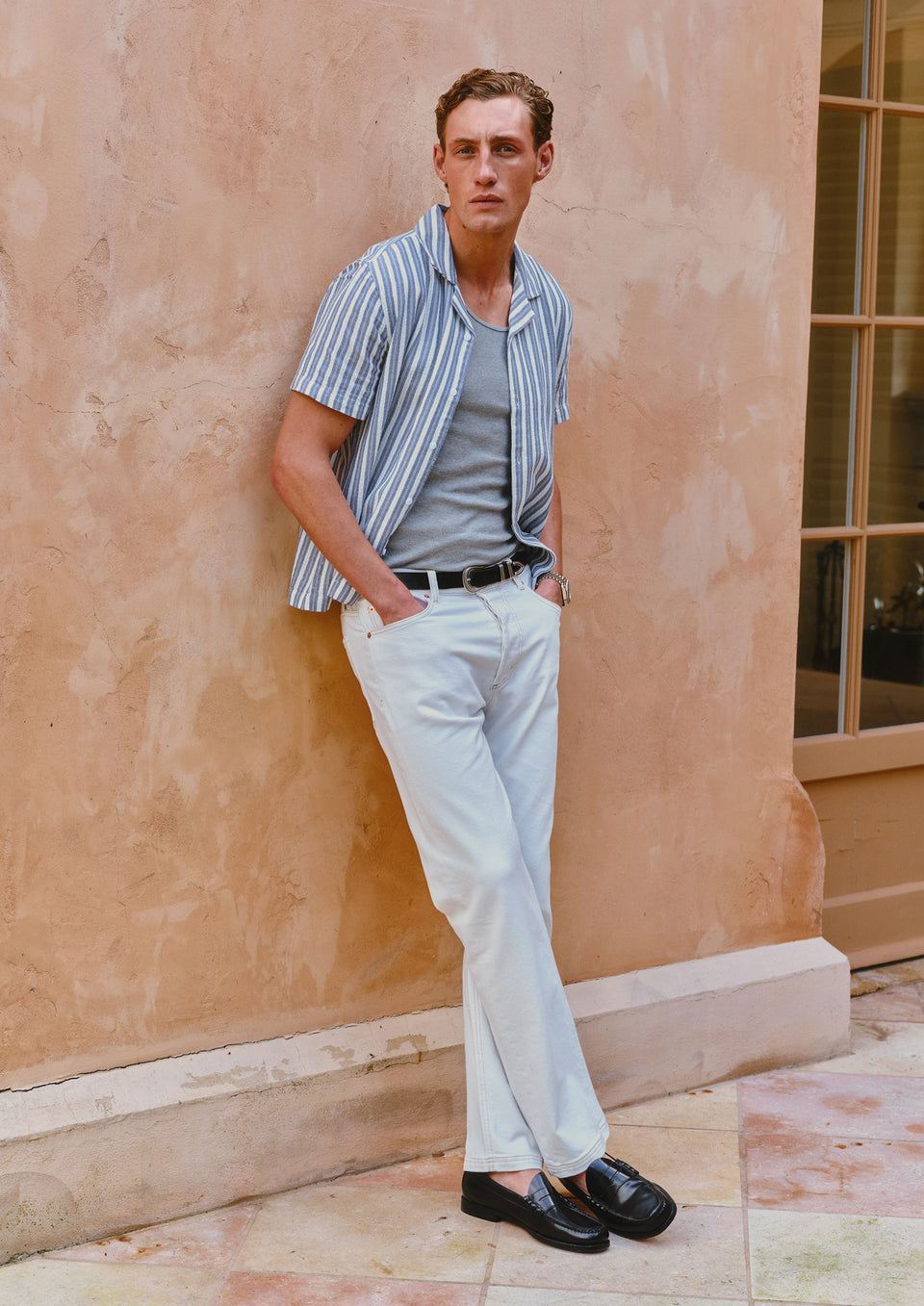 Man in striped shirt and light pants leaning against a beige wall.