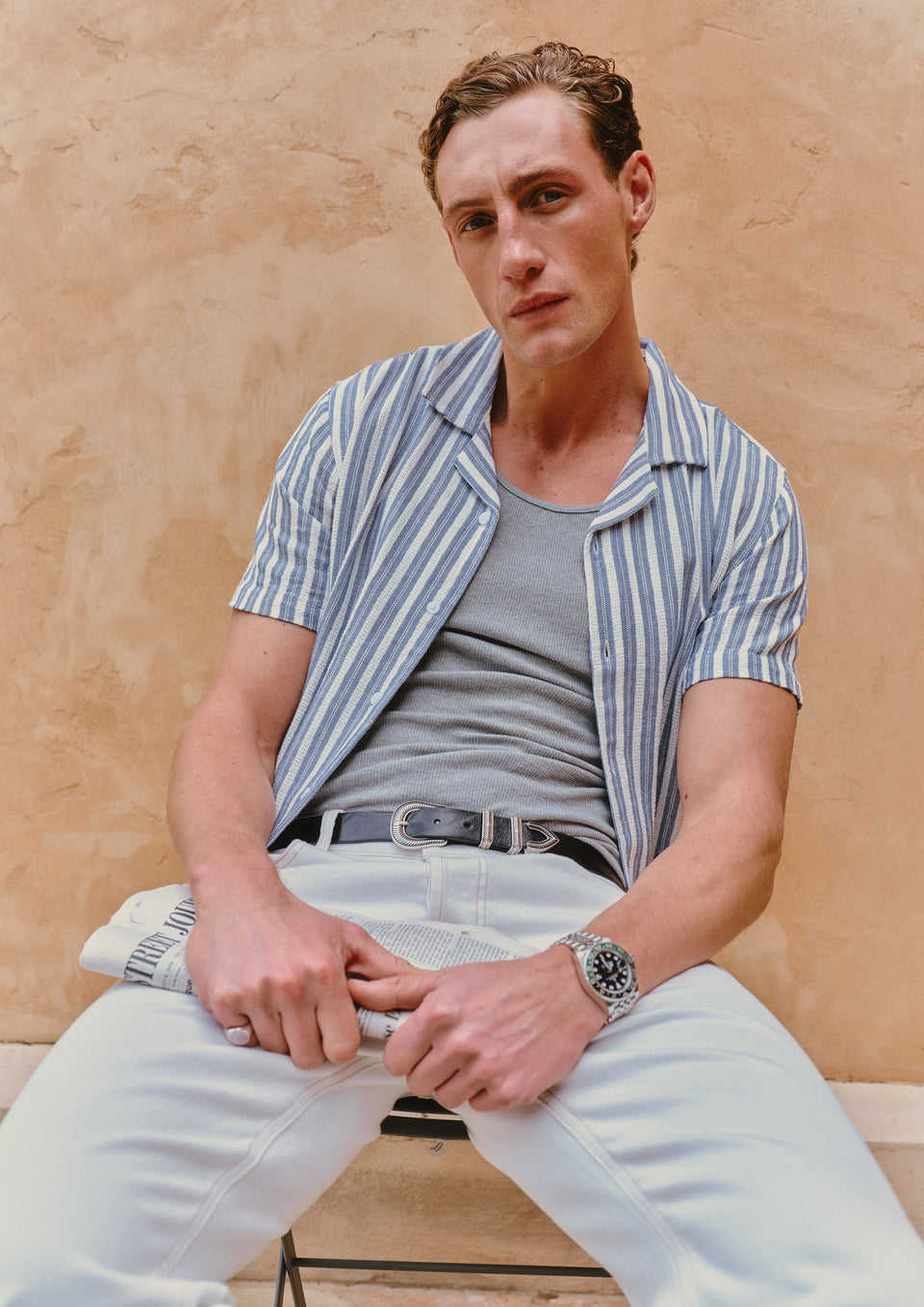 Man wearing a striped shirt over a gray t-shirt and light pants against a beige wall.