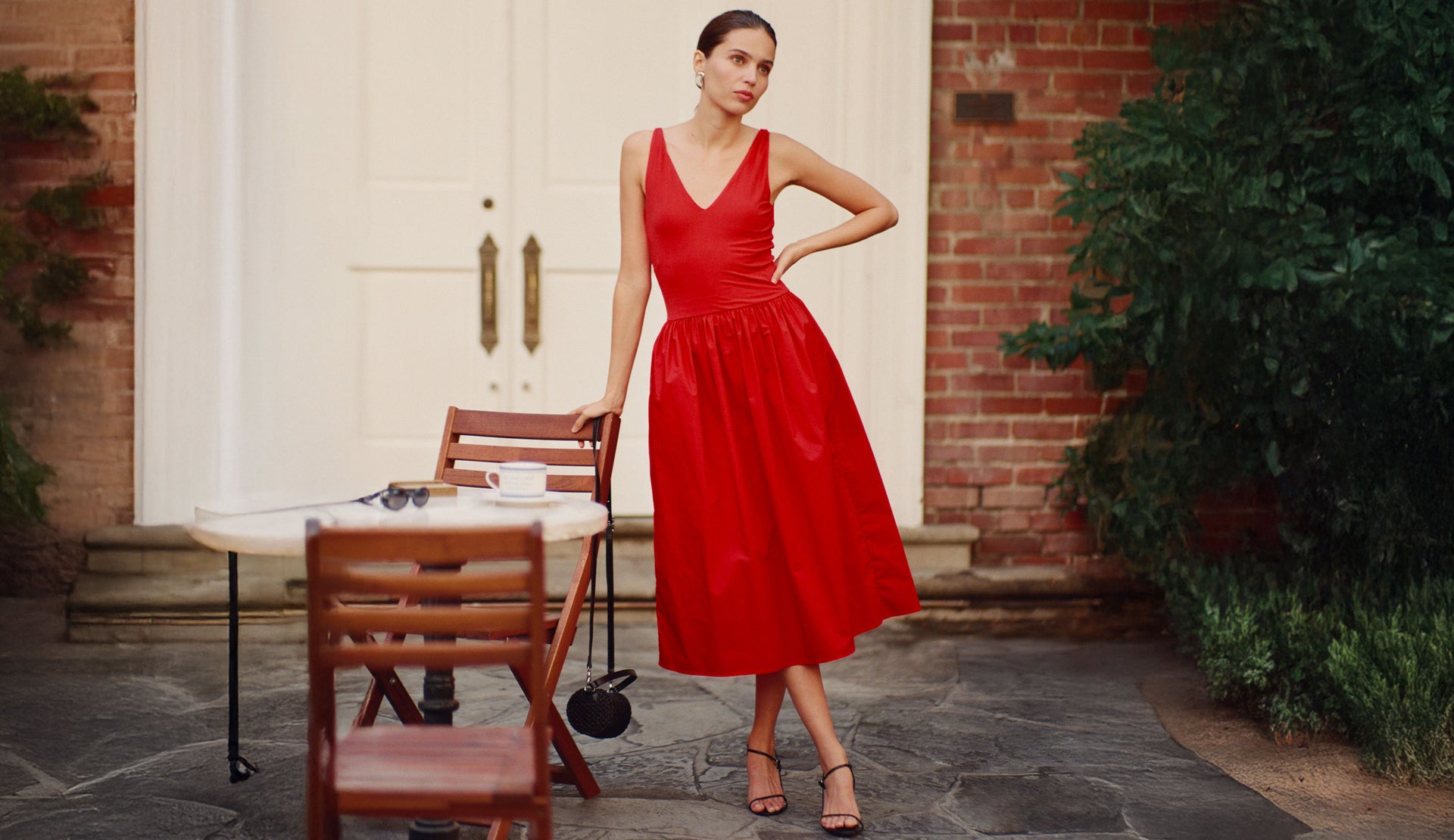 Woman in a red dress standing outdoors near a brick wall and table.