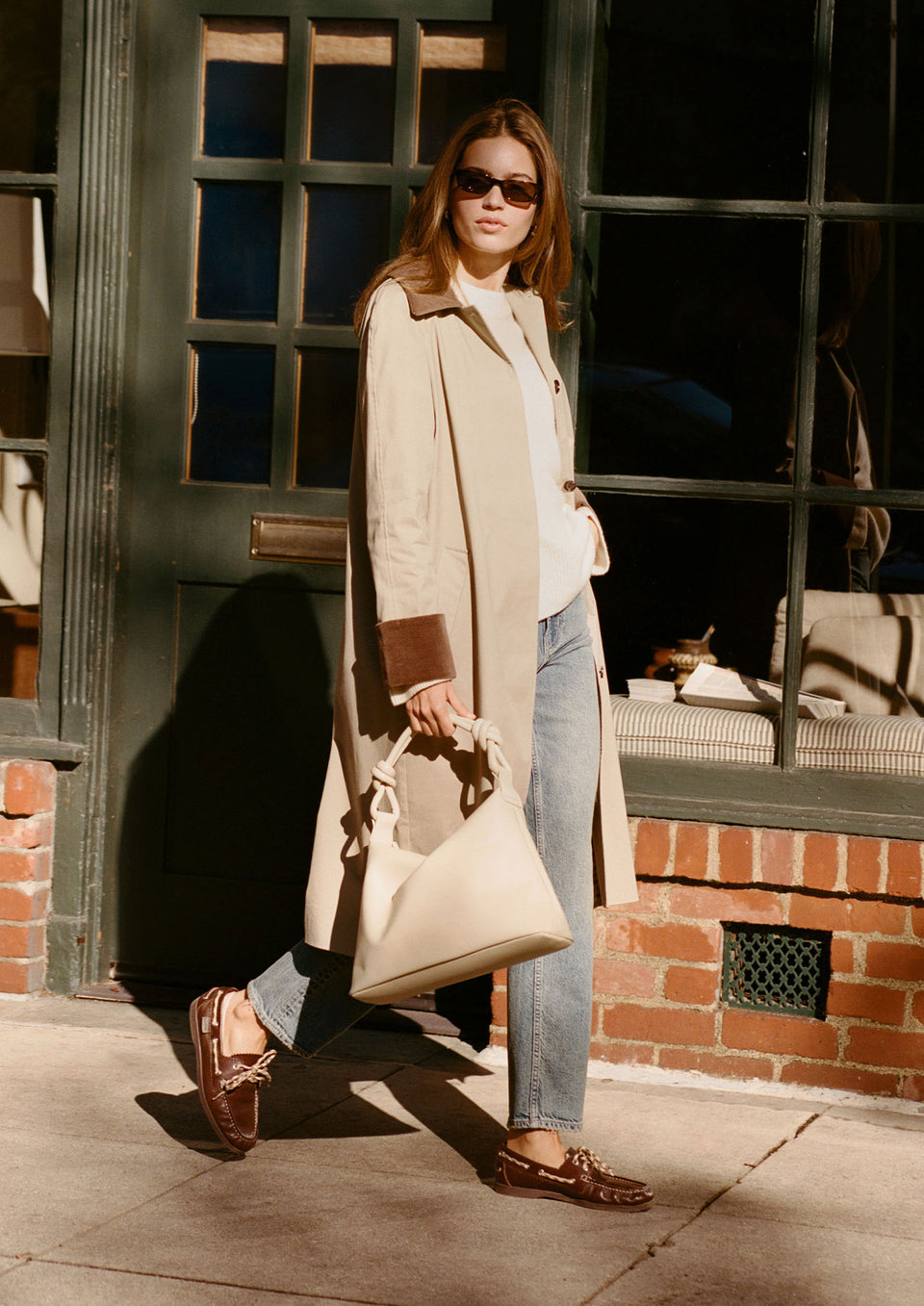 Woman in a beige coat and sunglasses holding a beige handbag.