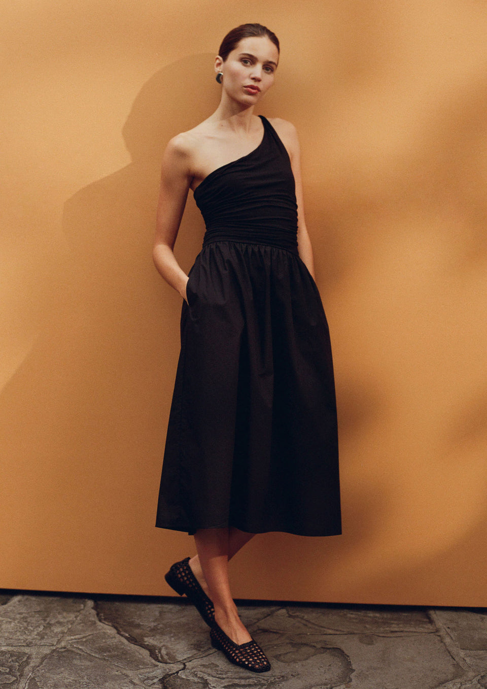 Woman wearing a black one-shoulder dress against an orange wall.
