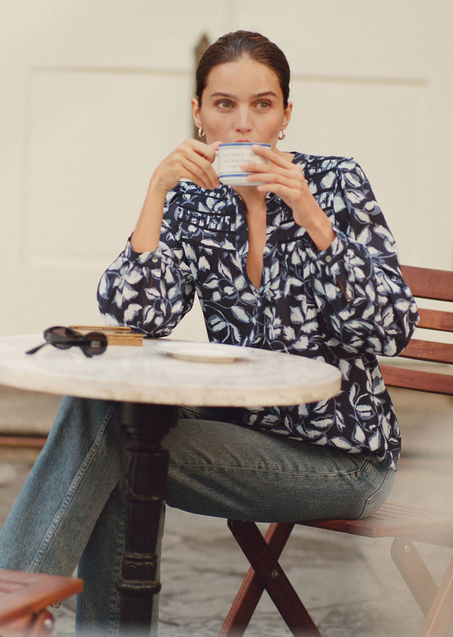 Woman sitting at a table outdoors, holding a cup.
