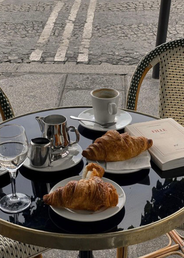 Breakfast setting with croissants, coffee, and a glass on a table outdoors.