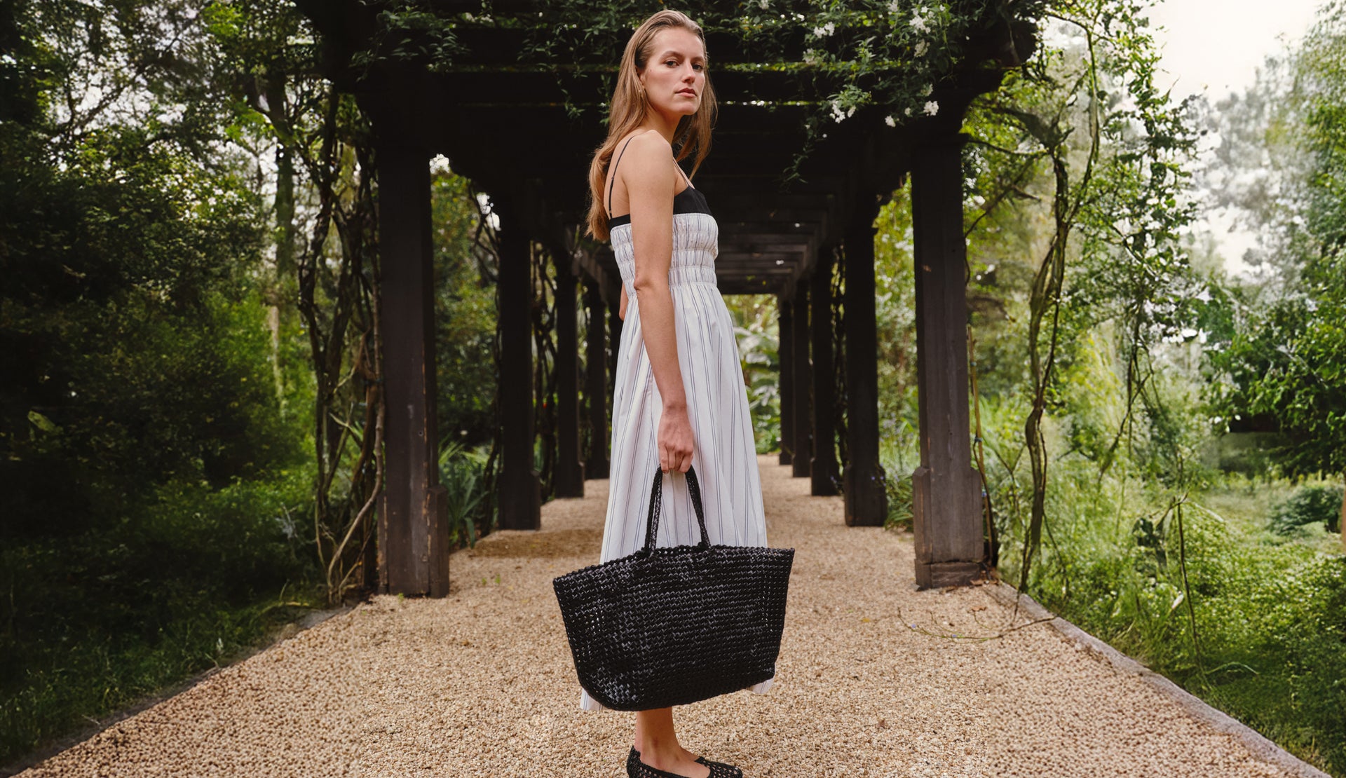 Woman in a white striped dress holding a black bag in a garden setting