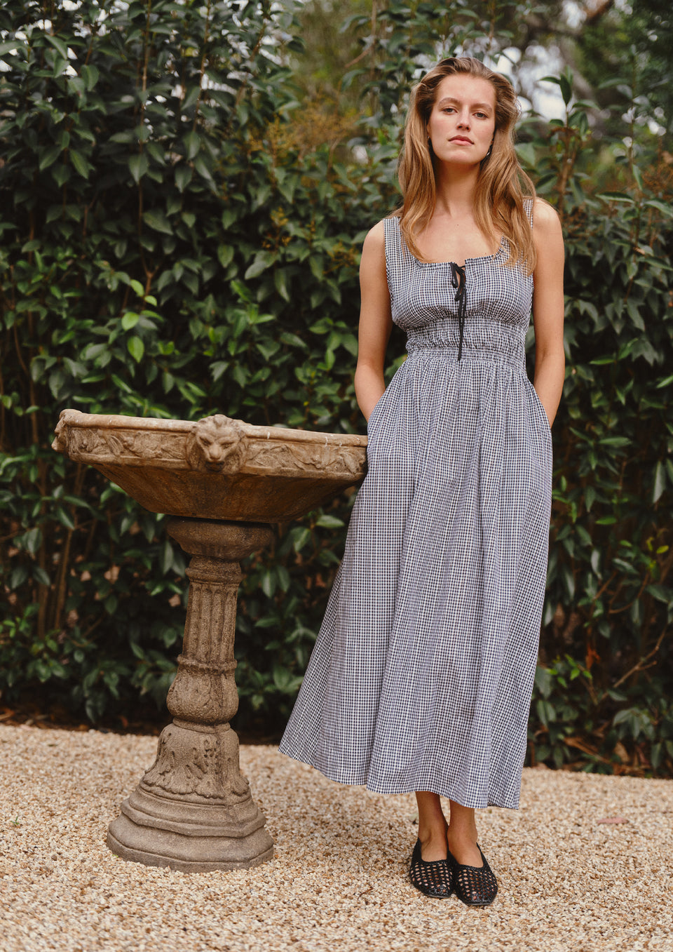 Woman in a checkered dress standing next to an old stone fountain with greenery in the background