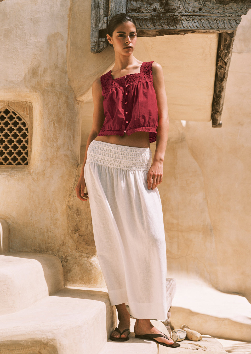 Woman wearing a red top and white pants standing in front of a textured wall.