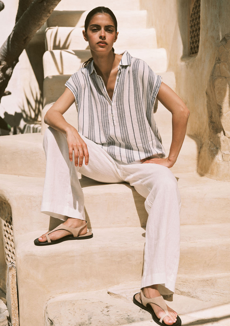 Woman sitting on stone steps wearing a striped shirt and white pants.