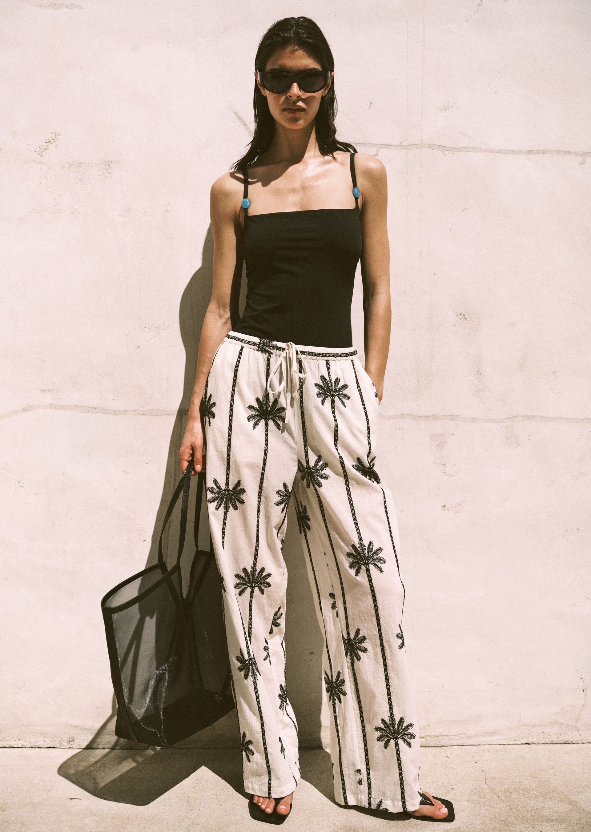 Woman wearing a black bathing suit and beige pants with black patterns against a light-colored wall.
